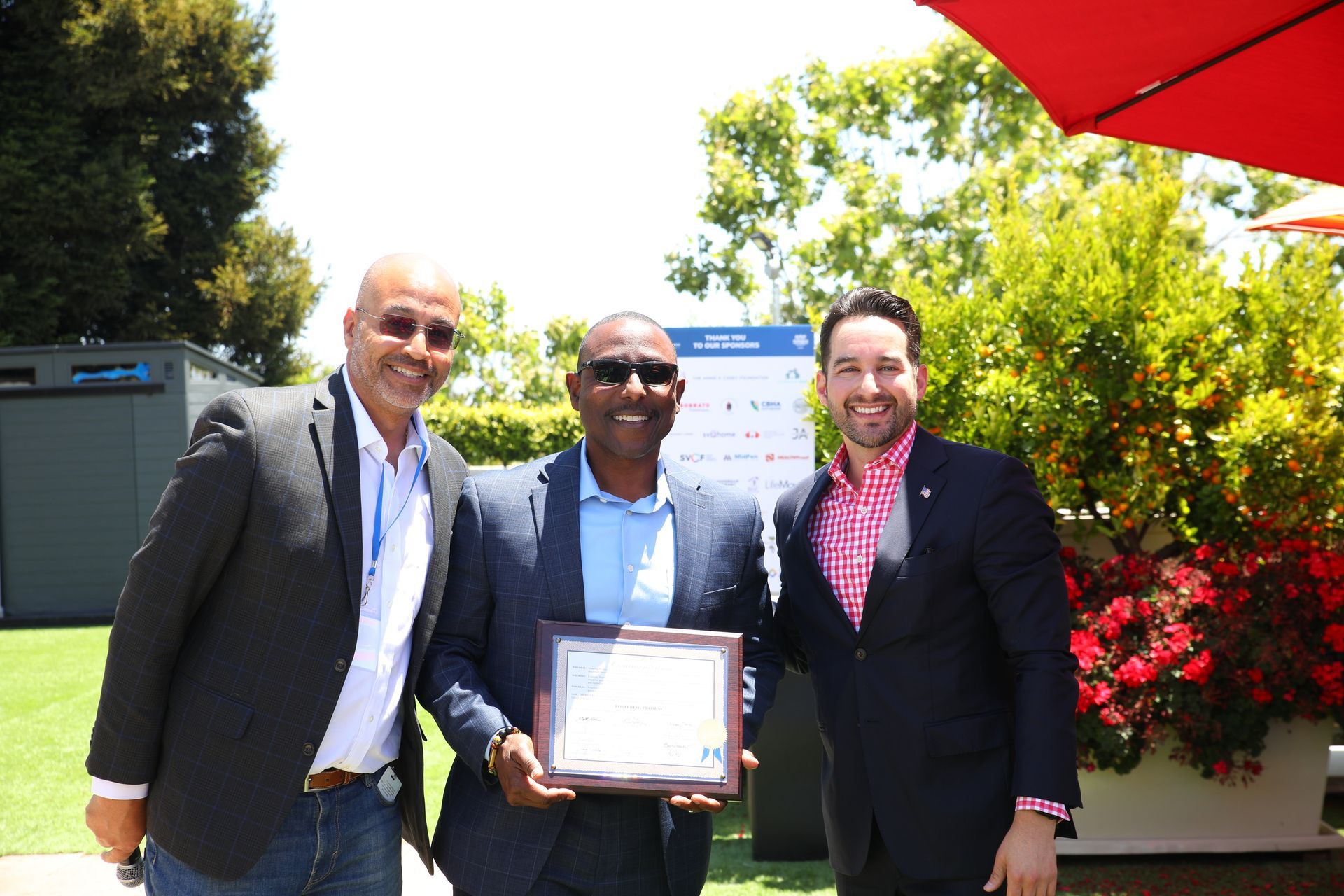 Three men in suits smiling outdoors, one holding a framed certificate.