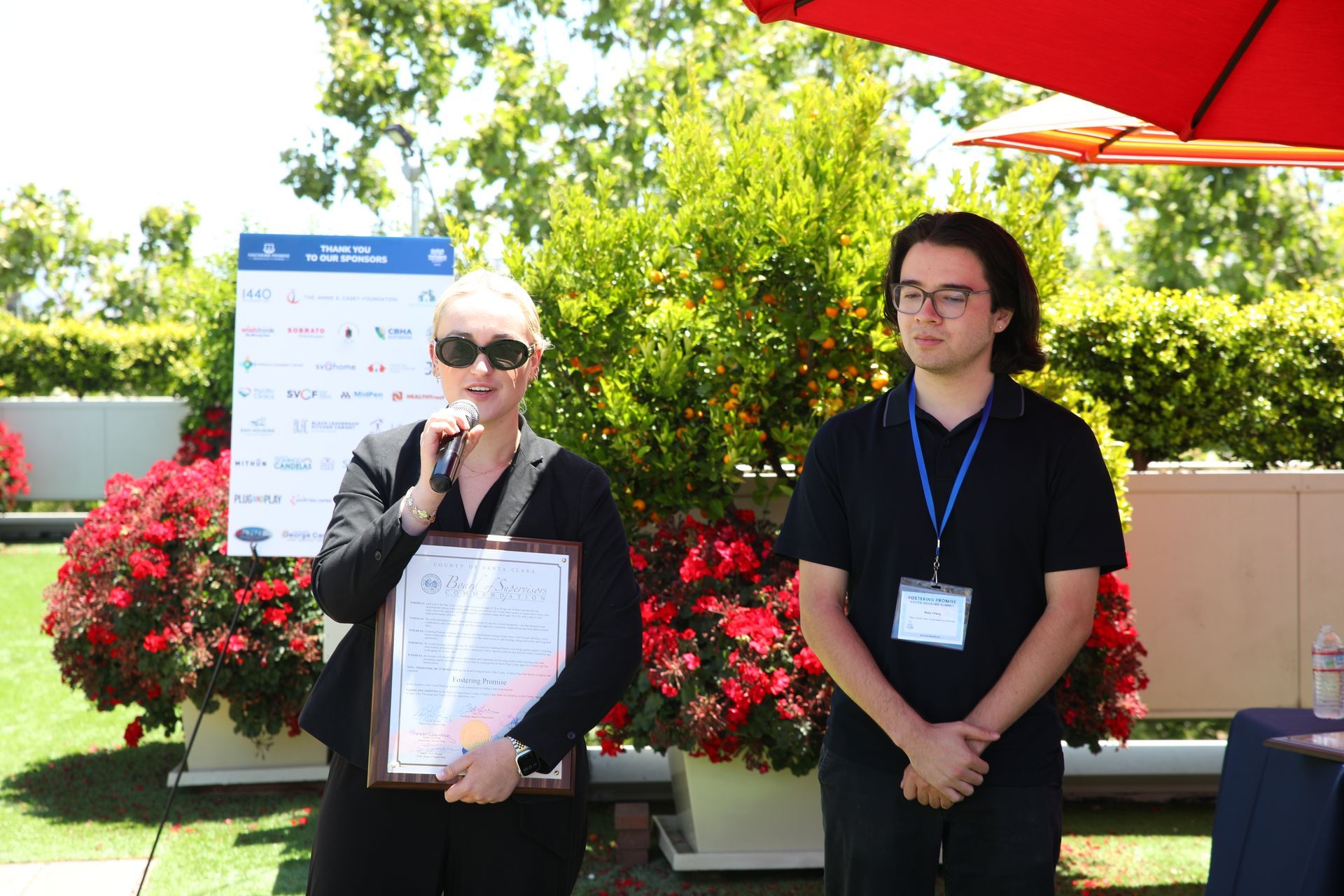 Two people at an outdoor event, one speaking into a microphone while holding a framed certificate.