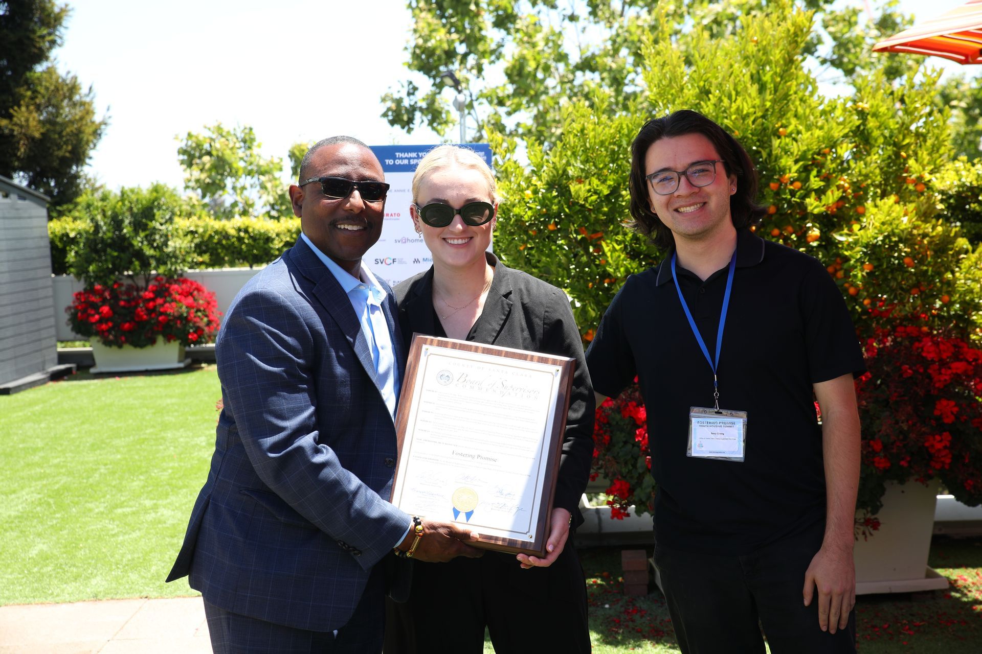 Three people holding a framed certificate outdoors in a sunny garden.