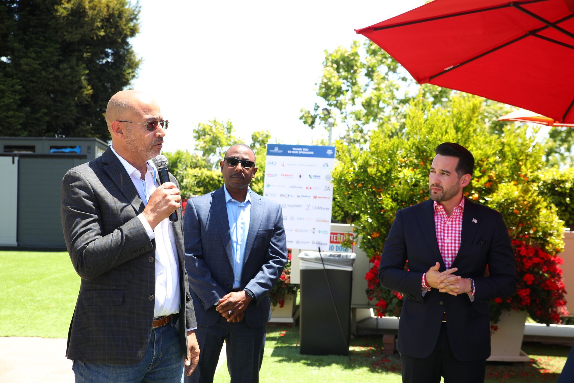 Three men in suits talking outdoors under a red umbrella at a sunny event