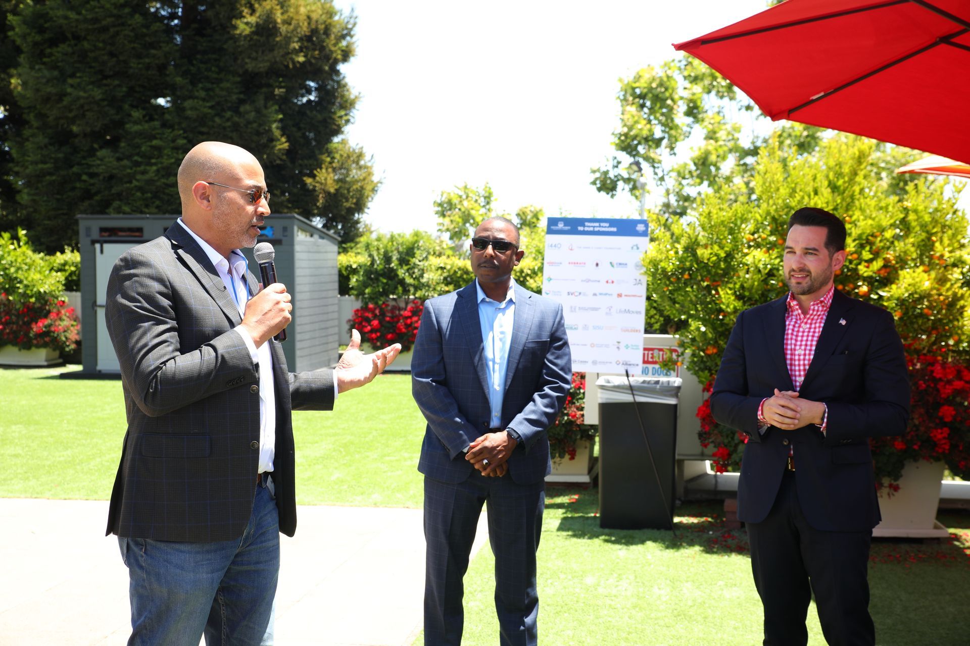 Three men stand outdoors at a speaking event, one holding a microphone under a red umbrella.