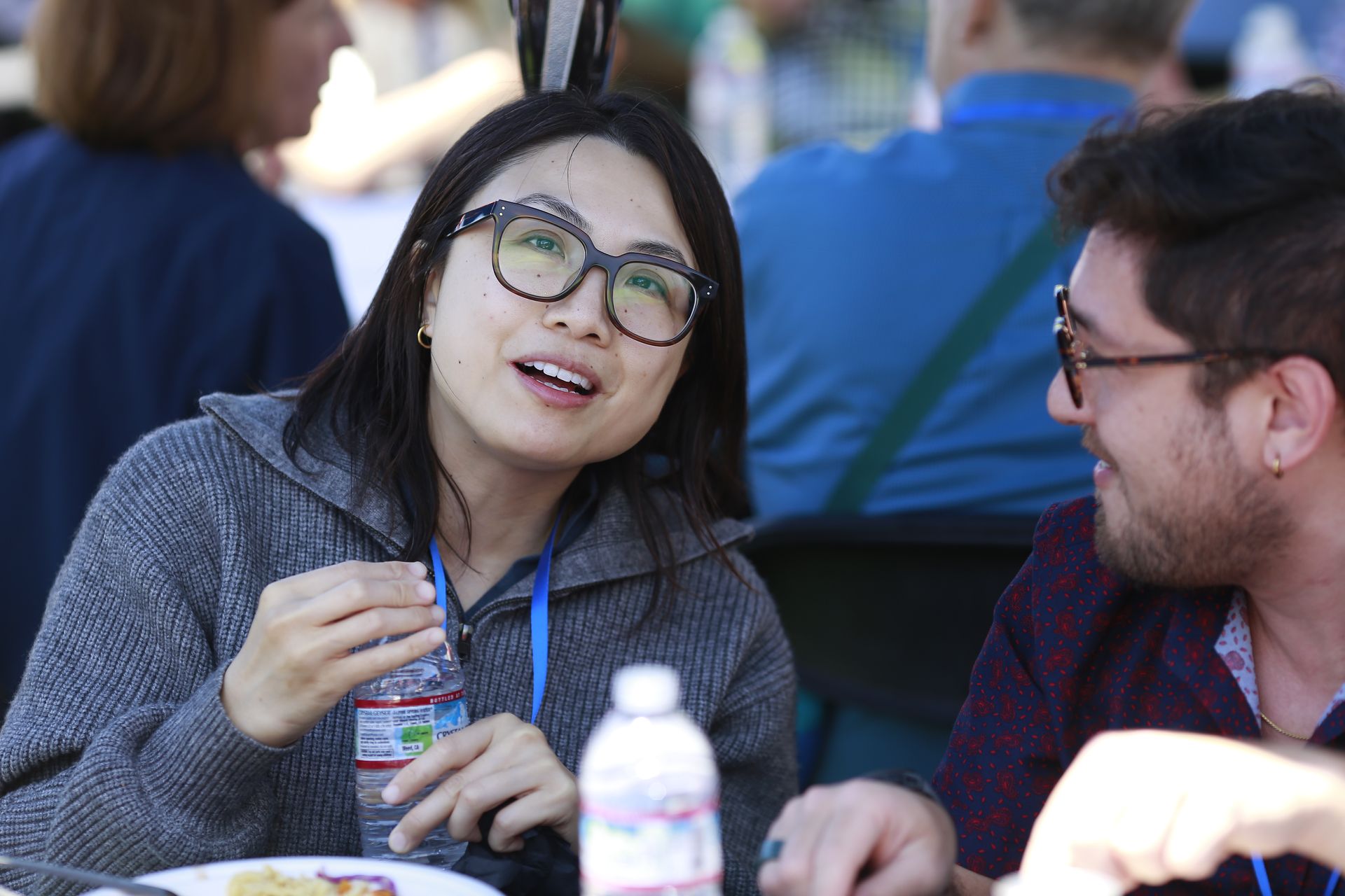 Two people talking at an outdoor table, one wearing glasses and holding a drink bottle.