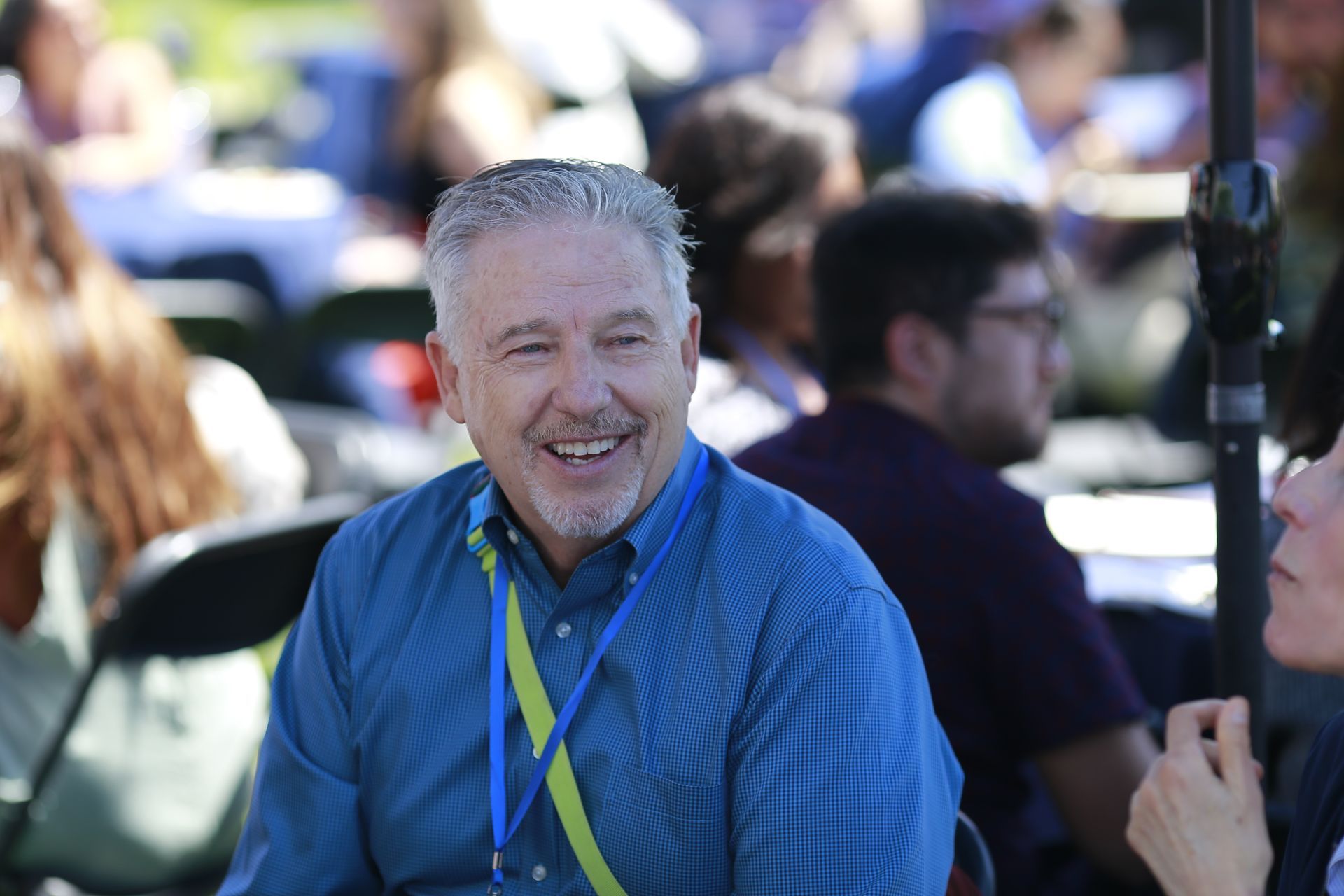 Smiling man in a blue shirt and lanyard at an outdoor event, with people blurred in the background