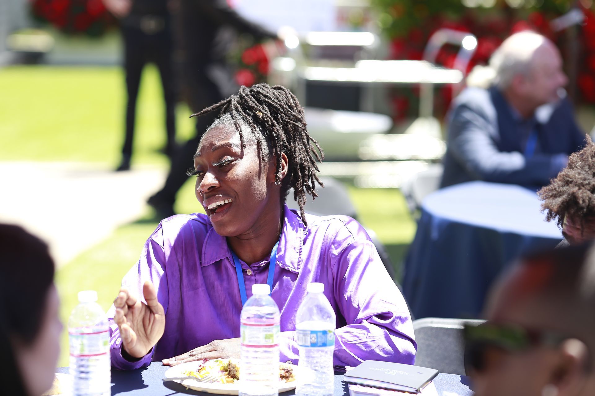 Person in a purple shirt speaking at an outdoor table with drinks and plates during a sunny gathering