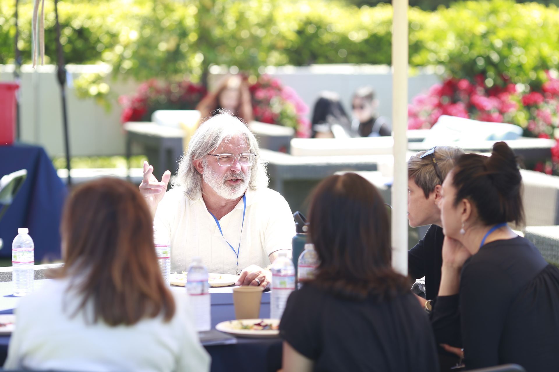 Outdoor panel discussion with an older speaker at a table, audience seated in the foreground.