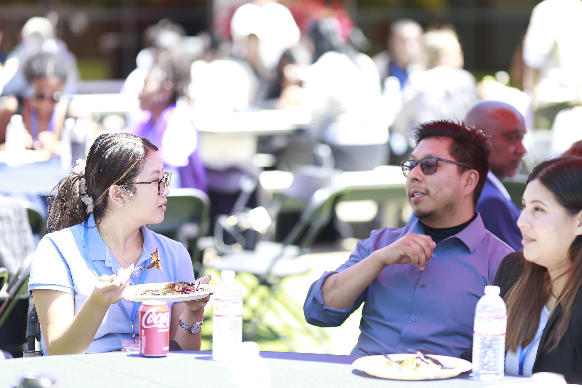 Three people talking at an outdoor picnic table with drinks in a sunny park
