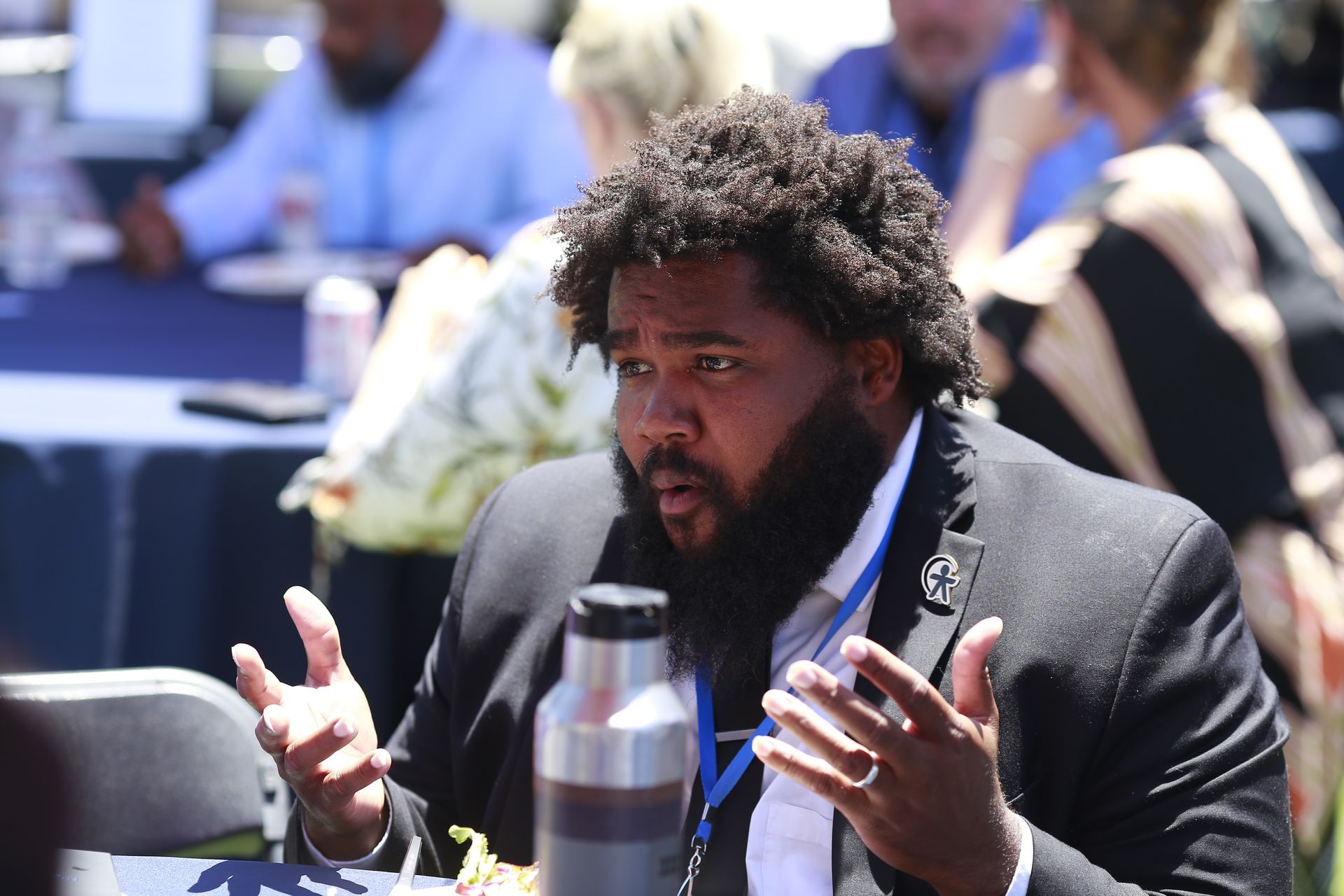 Man in a suit speaking animatedly at an outdoor table with a water bottle in the foreground