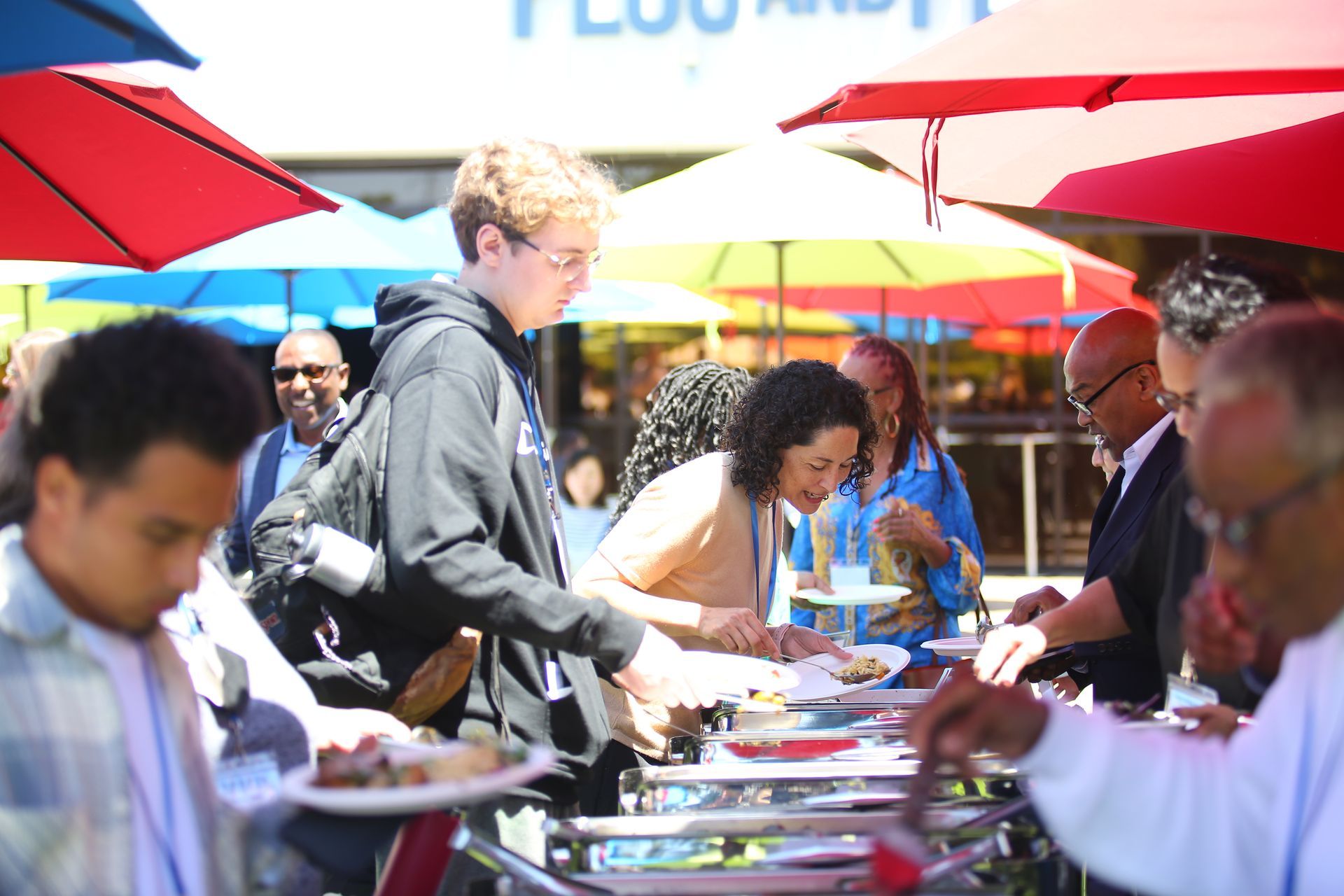 People serving food at an outdoor buffet under red umbrellas in bright sunlight
