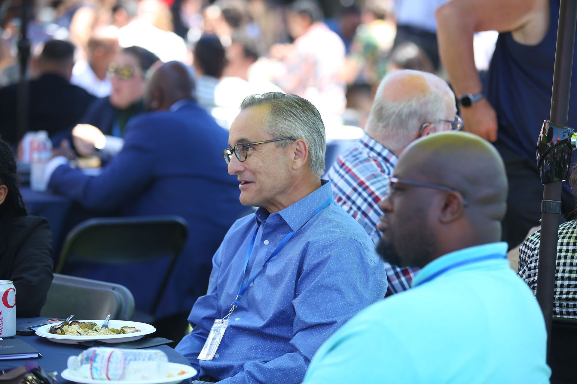 Two men seated at an outdoor event table, one in a blue shirt and one in a light blue shirt, with plates nearby