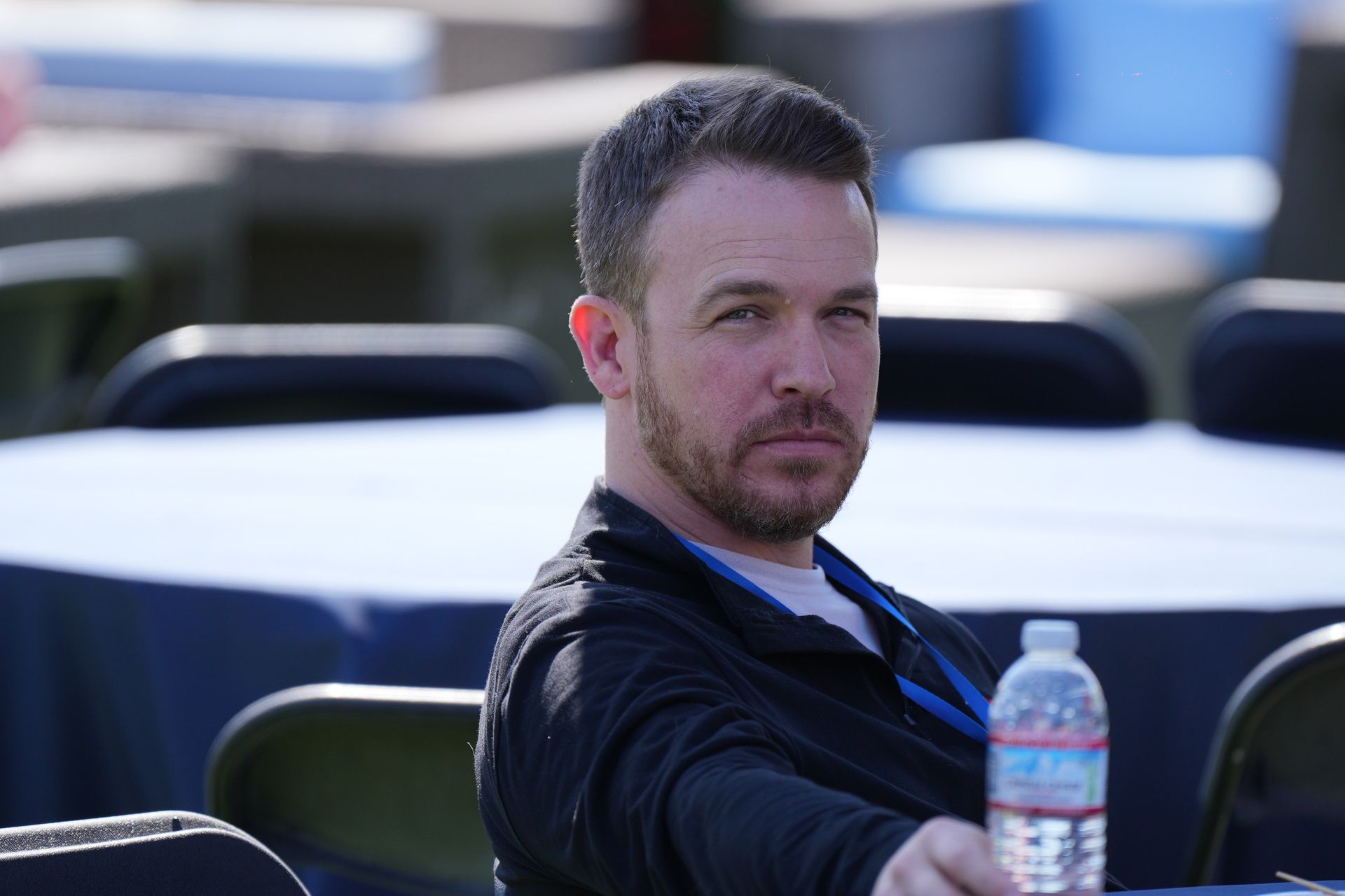 Man seated outdoors at a table, wearing a dark shirt and looking toward the camera, with a water bottle in front.