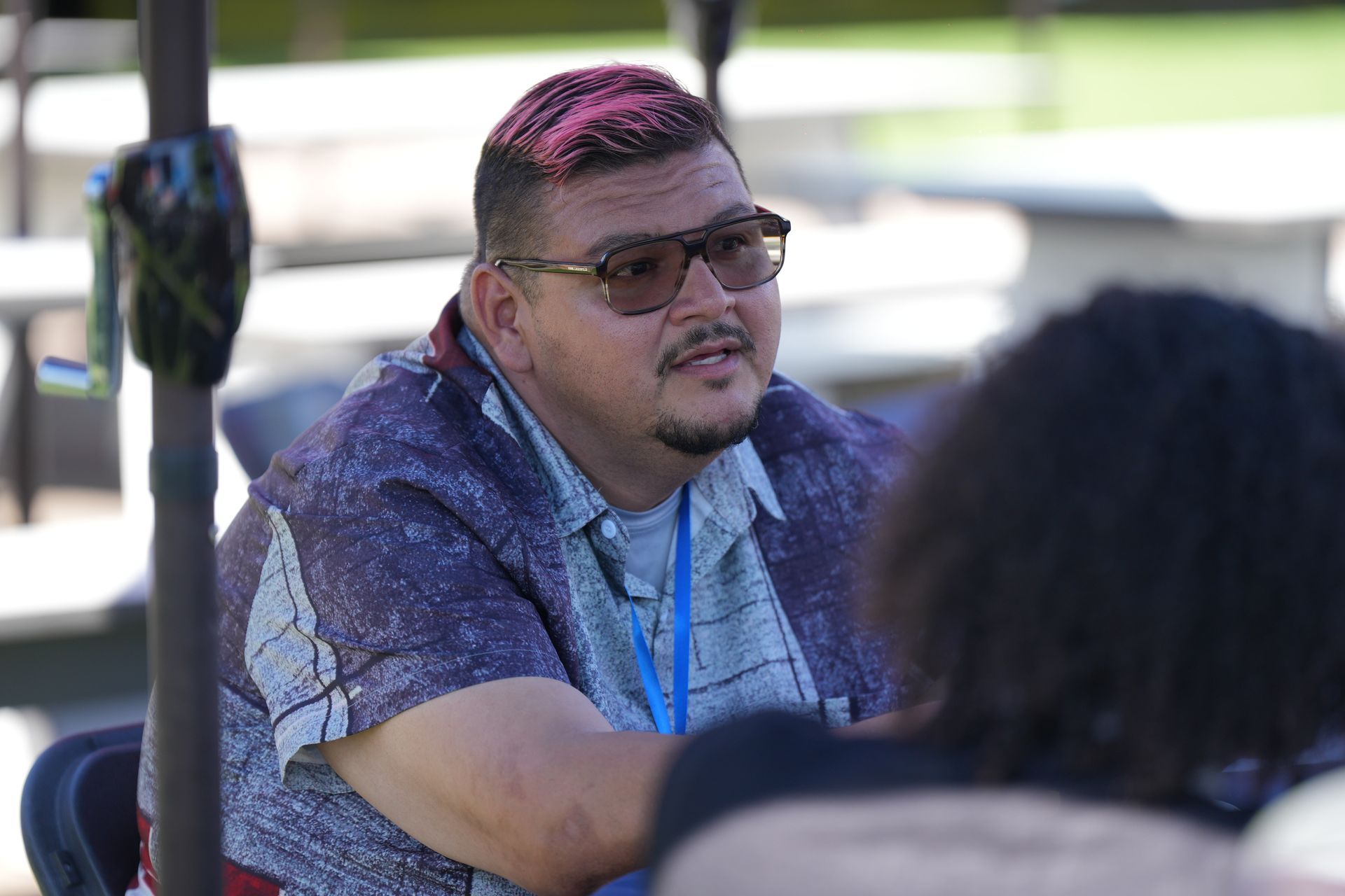 Man with pink-topped hair and glasses talking outdoors at a table, with another person in the foreground