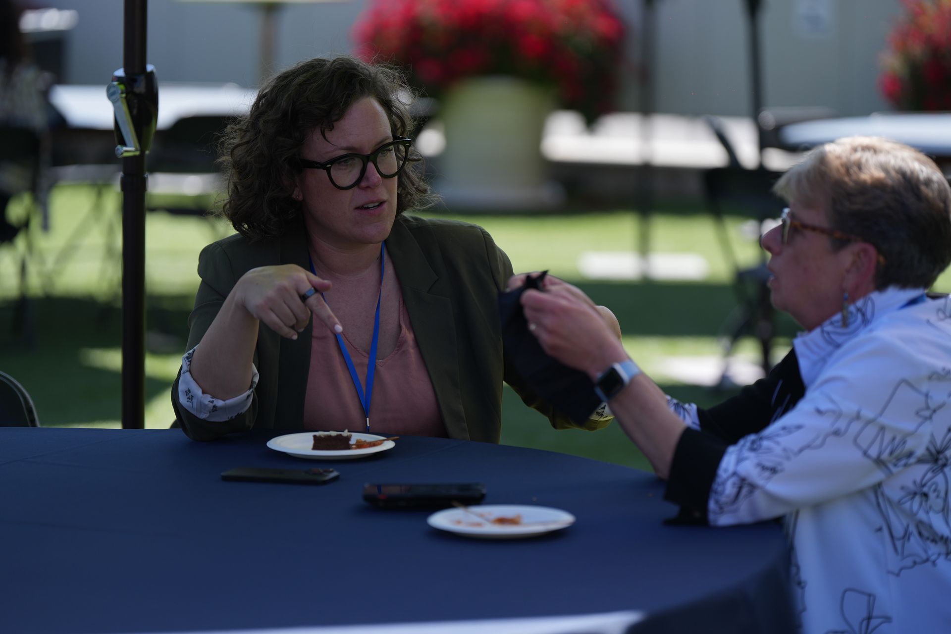 Two people talking at a round table outdoors, with plates and drinks in a sunny garden setting.
