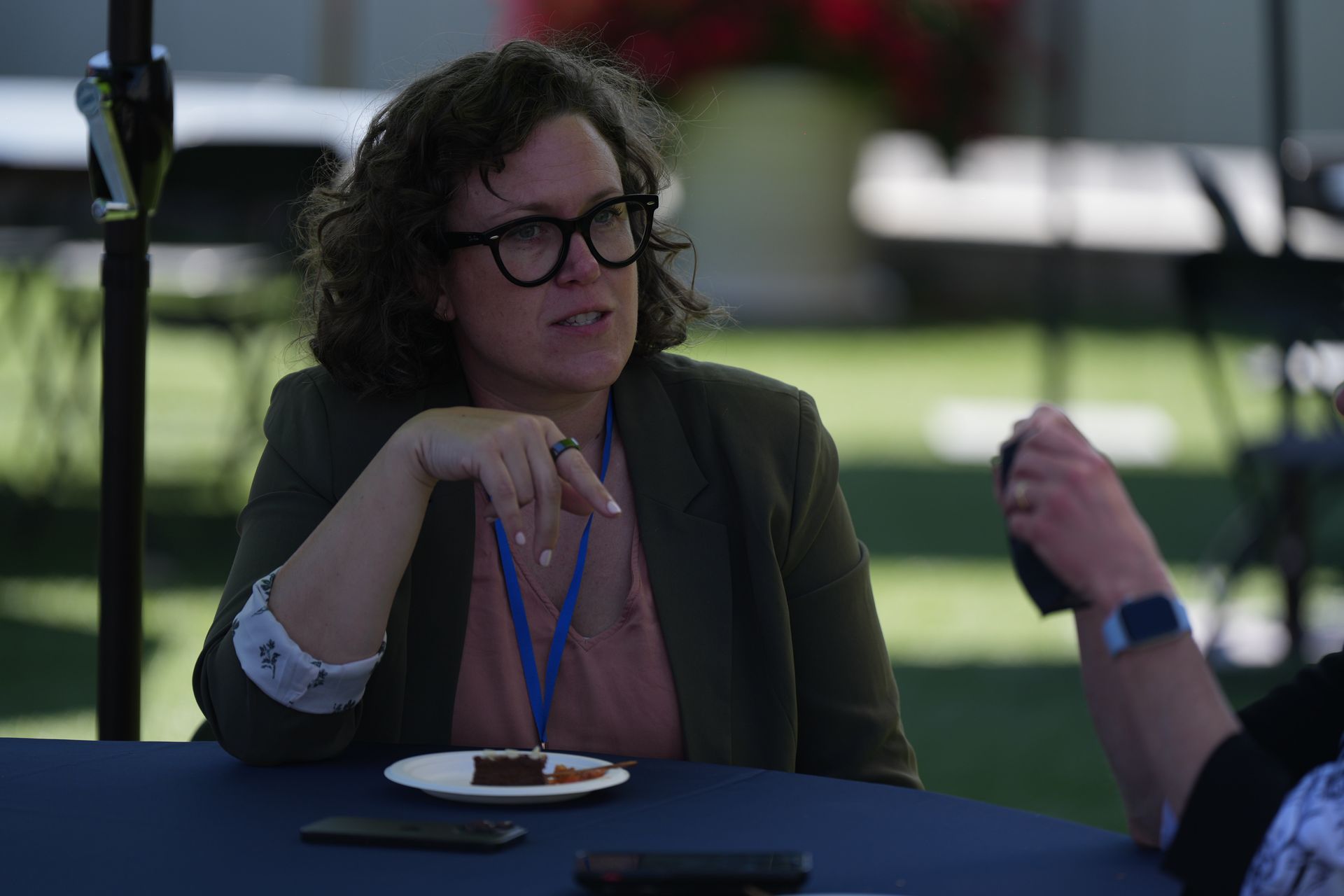Woman with glasses seated at a table outdoors, gesturing during a conversation, with a plate and cup nearby