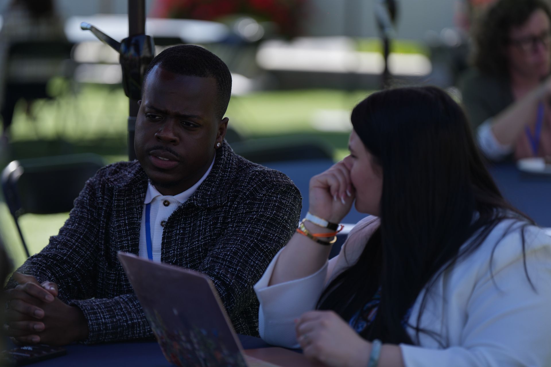 Two people talking at an outdoor table with a laptop in a shaded park-like setting