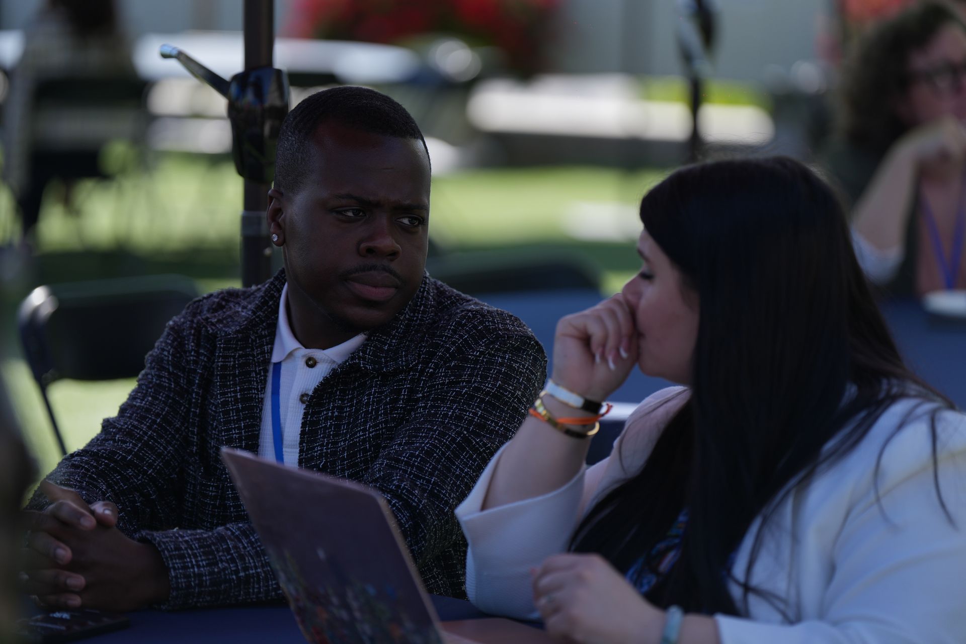 Two people seated outdoors, talking beside an open laptop under a shaded canopy.