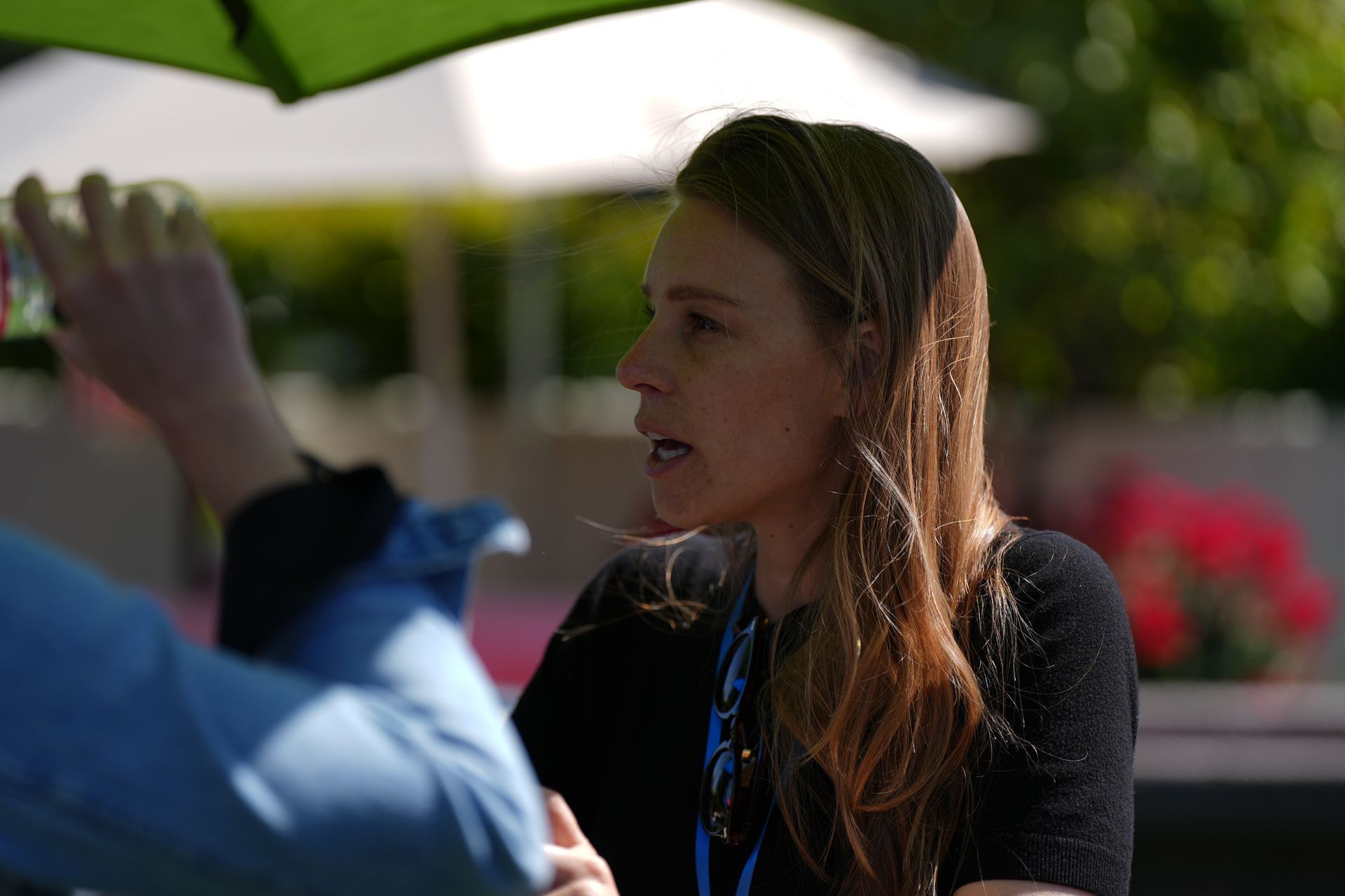 Woman speaking outdoors under a green umbrella, with a blurred person in the foreground.