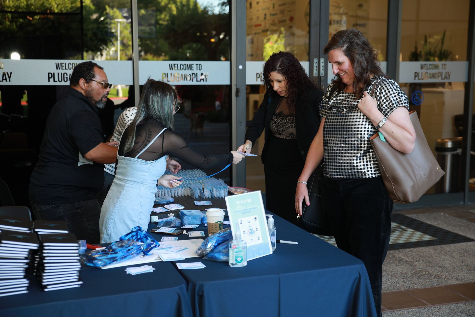 People at a blue table with brochures, one handing items to two attendees outside a building