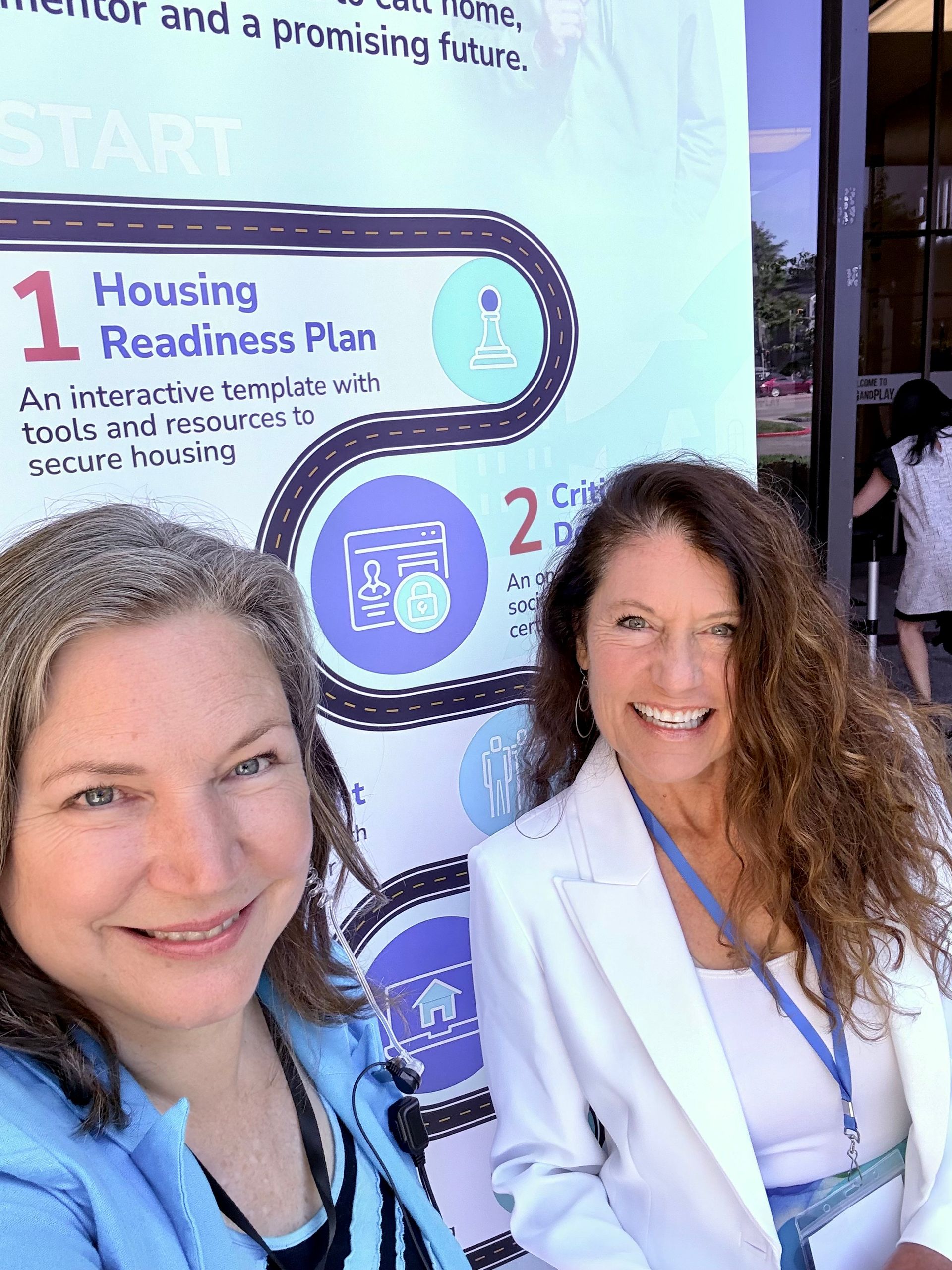 Two women smiling beside a “Housing Readiness Plan” poster at an event booth