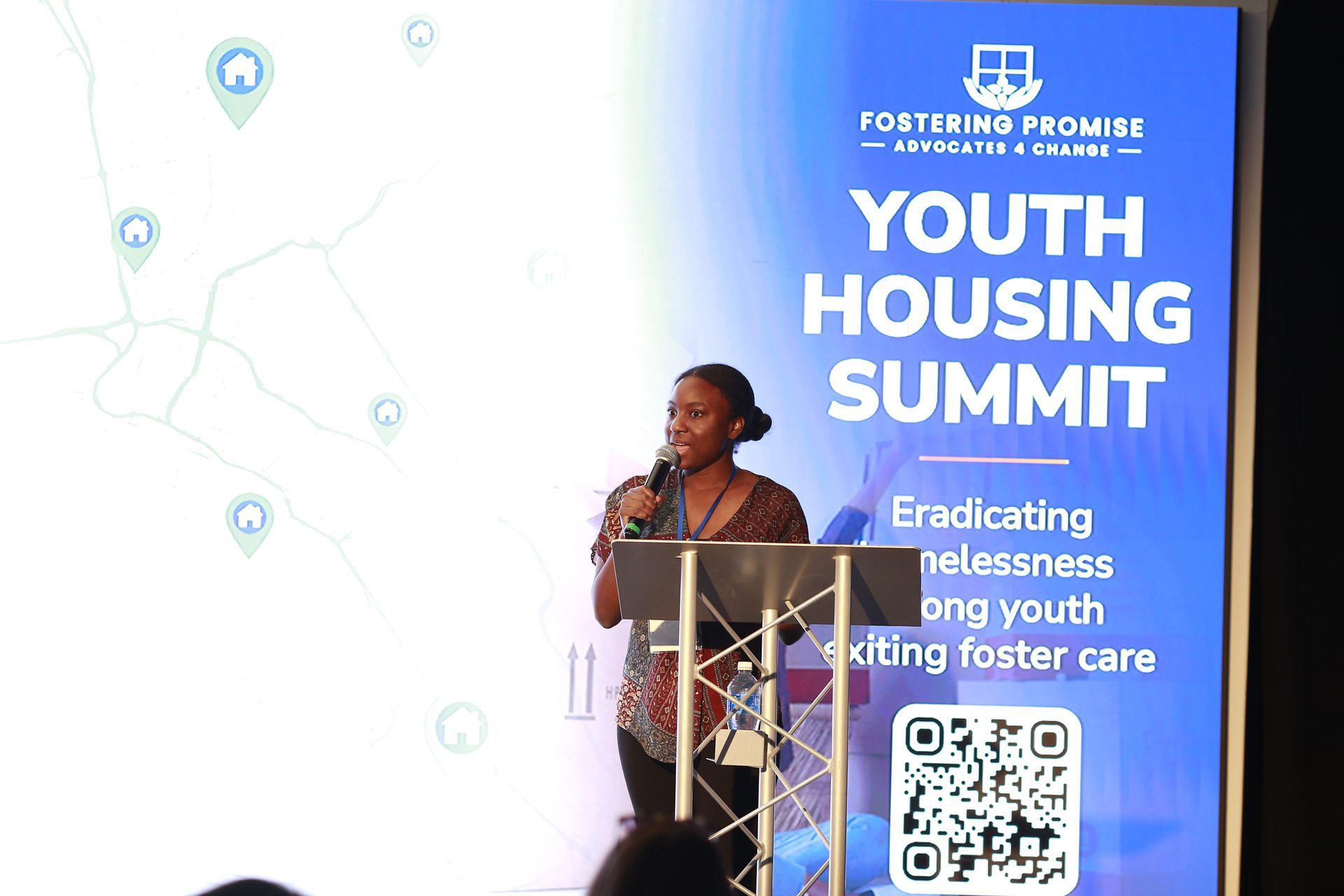 Speaker at podium during Youth Housing Summit, standing beside a blue event banner and QR code.