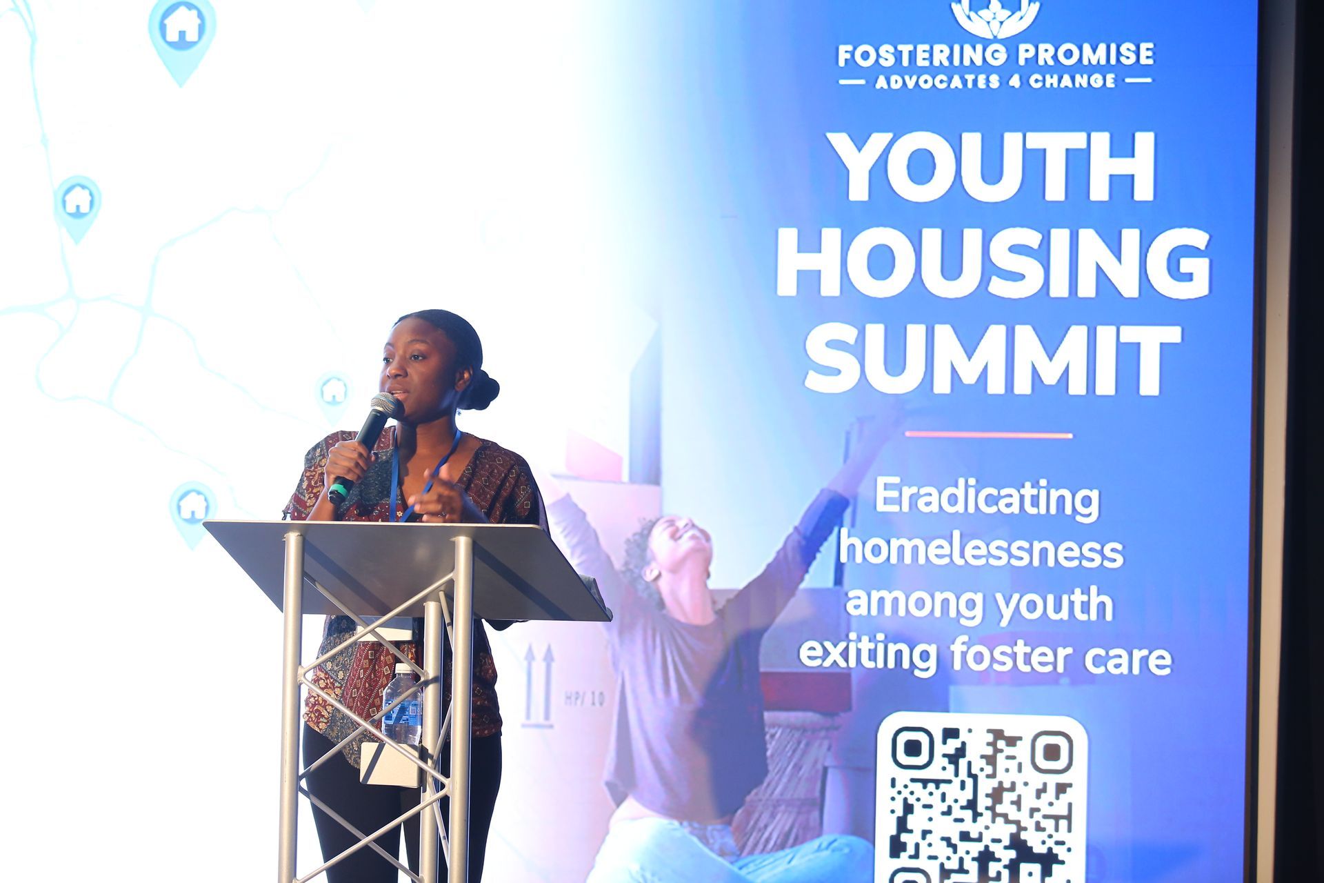 Speaker at podium beside “Youth Housing Summit” banner and QR code, discussing homelessness and foster care.