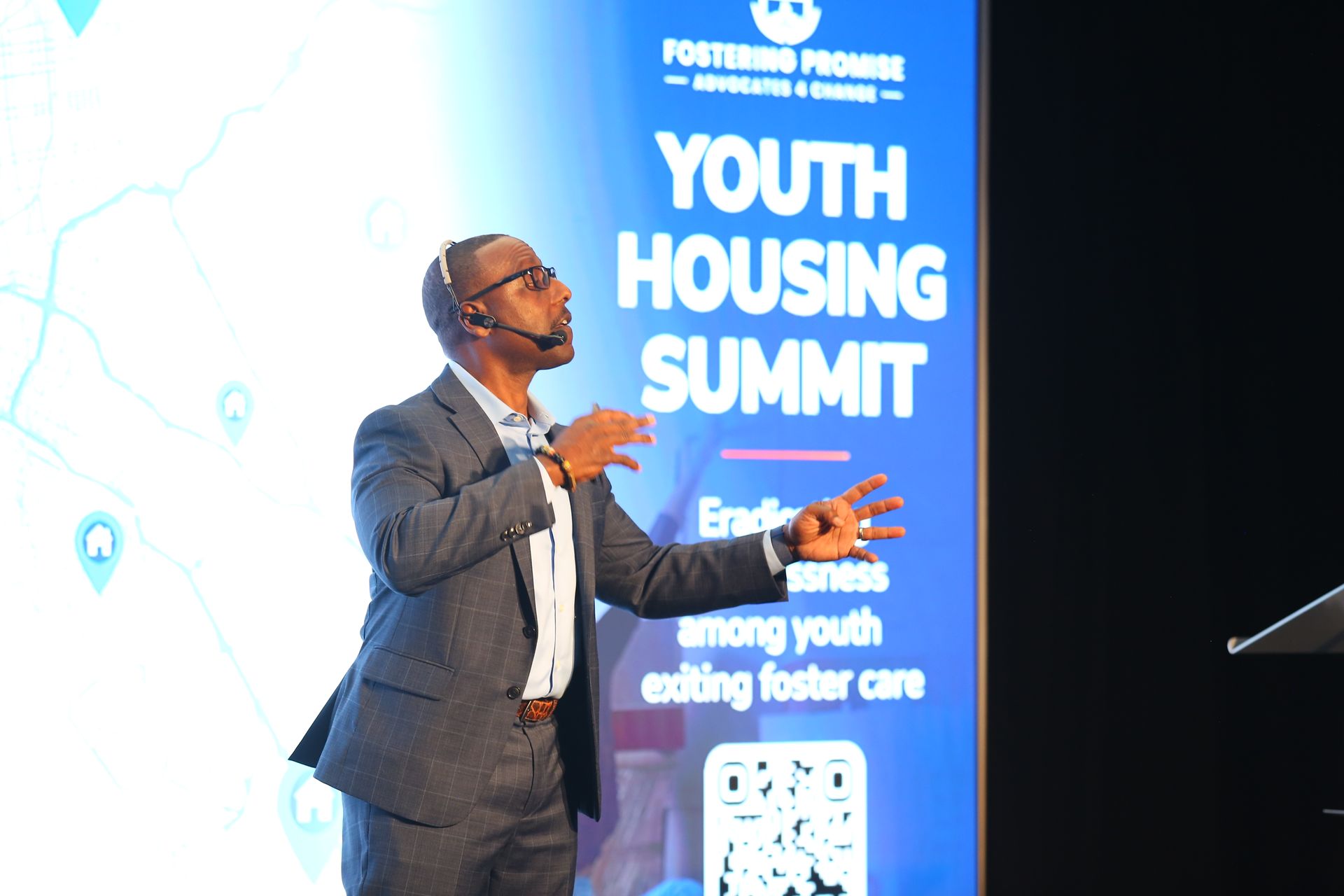 Speaker on stage beside a blue “Youth Housing Summit” banner, gesturing during a presentation