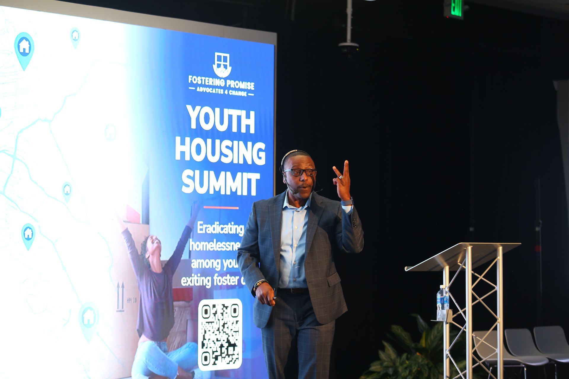 Speaker onstage at Youth Housing Summit beside blue event banner and podium, gesturing while presenting