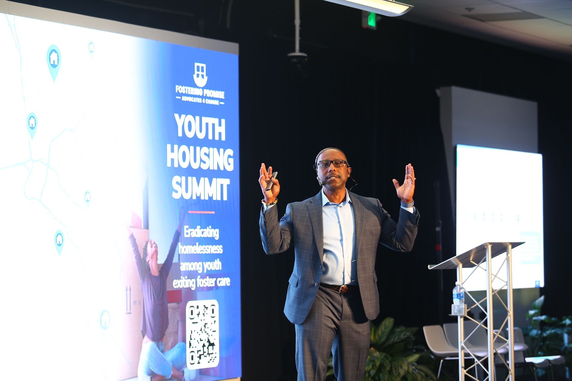 Speaker presenting at a Youth Housing Summit beside a blue event banner and podium on a stage
