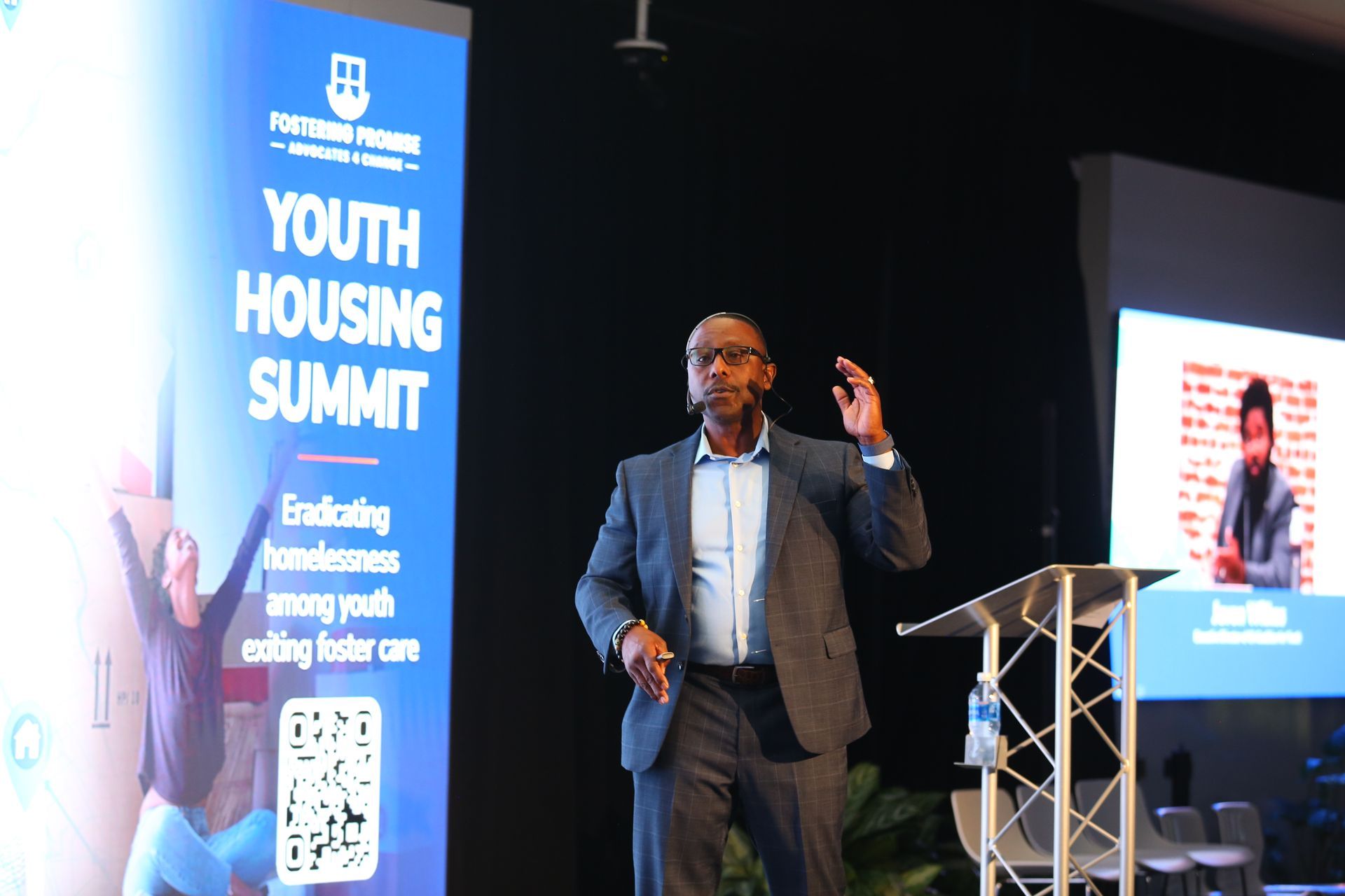 Speaker on stage at Youth Housing Summit, gesturing beside a blue event banner and podium