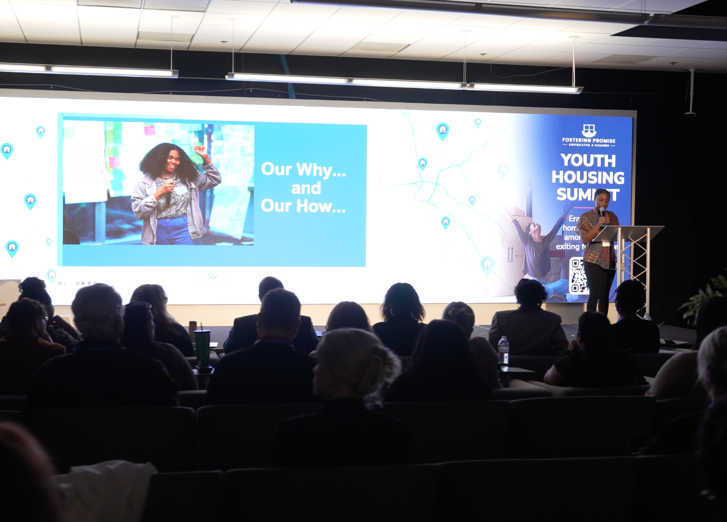 Conference stage with large screen reading “Our Why… and Our Now…” and a speaker at a podium, audience silhouetted in front.