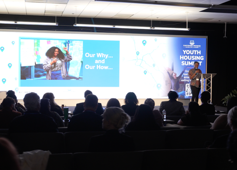 Speaker on stage at conference with large screen reading “Our Why… and Our Now…” and audience seated in foreground