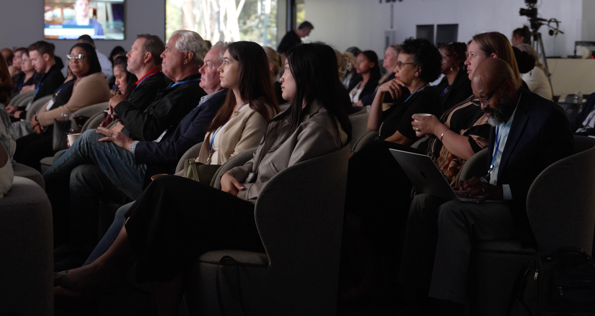 Audience seated in a dark conference room, watching a presentation and clapping