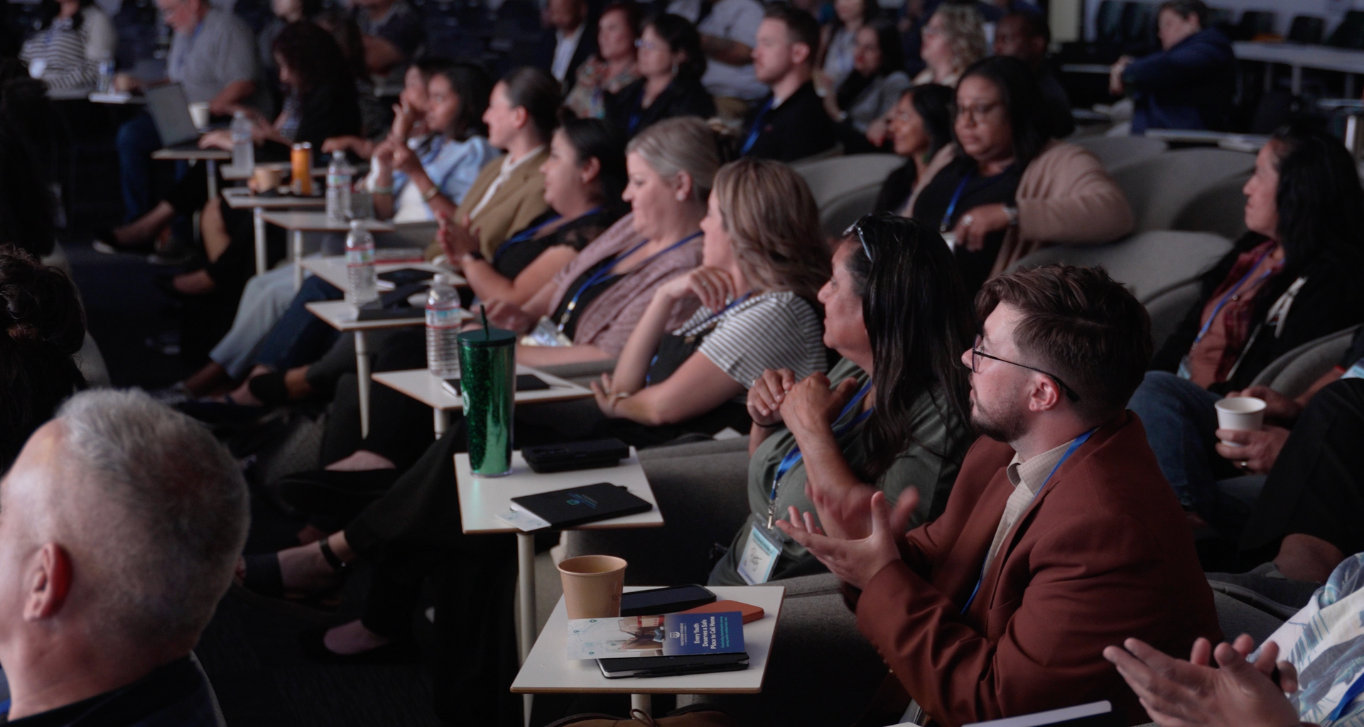 Audience seated at tables, applauding at a dimly lit conference or presentation