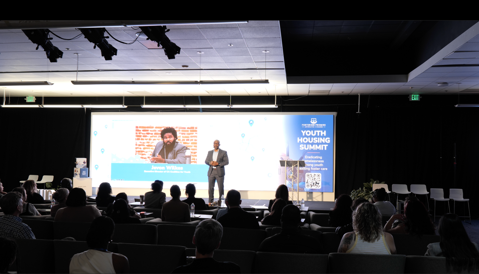 Conference presentation on a stage with two speakers, large slide screen, and seated audience in a dark auditorium