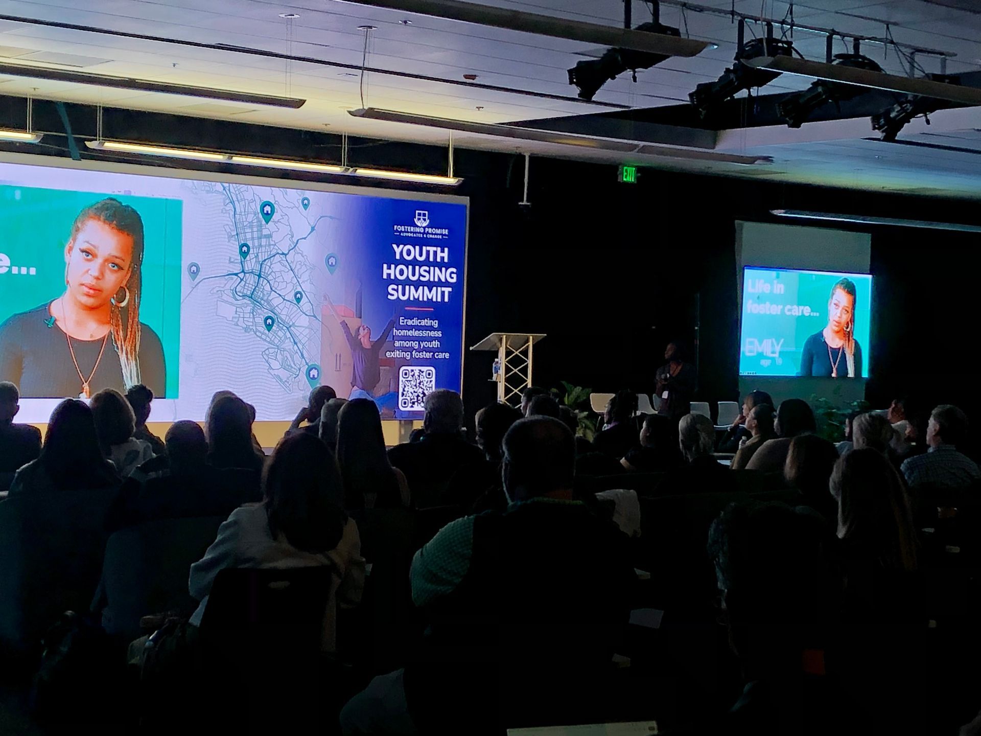 Conference audience watching a speaker on a large stage with blue event screens and a podium