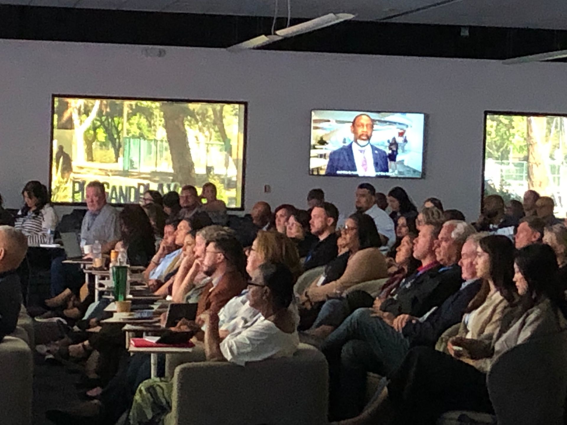 Audience seated in a dim conference room watching a presentation on wall-mounted screens