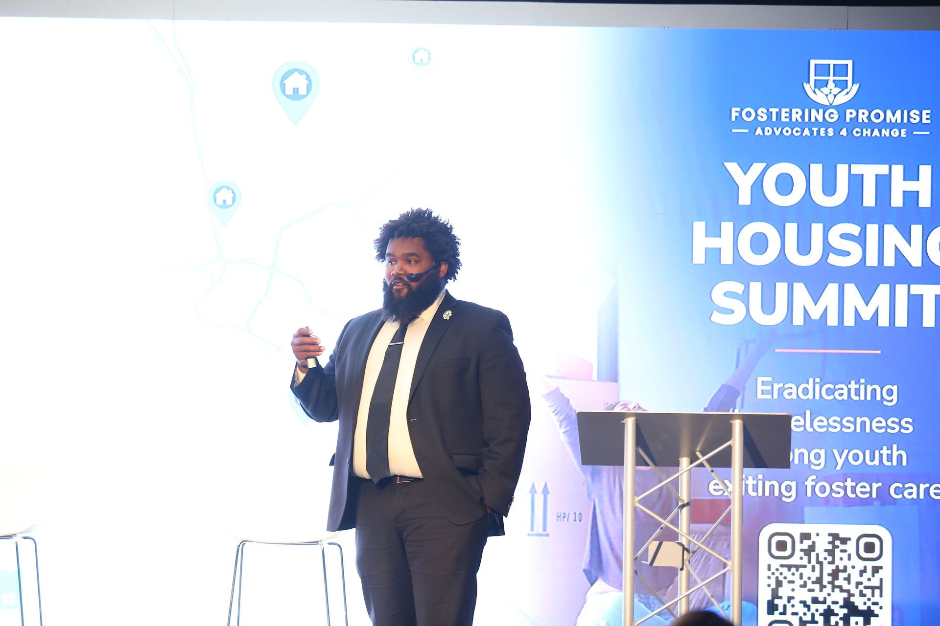 Speaker on stage at Youth Housing Summit beside a blue event banner and podium