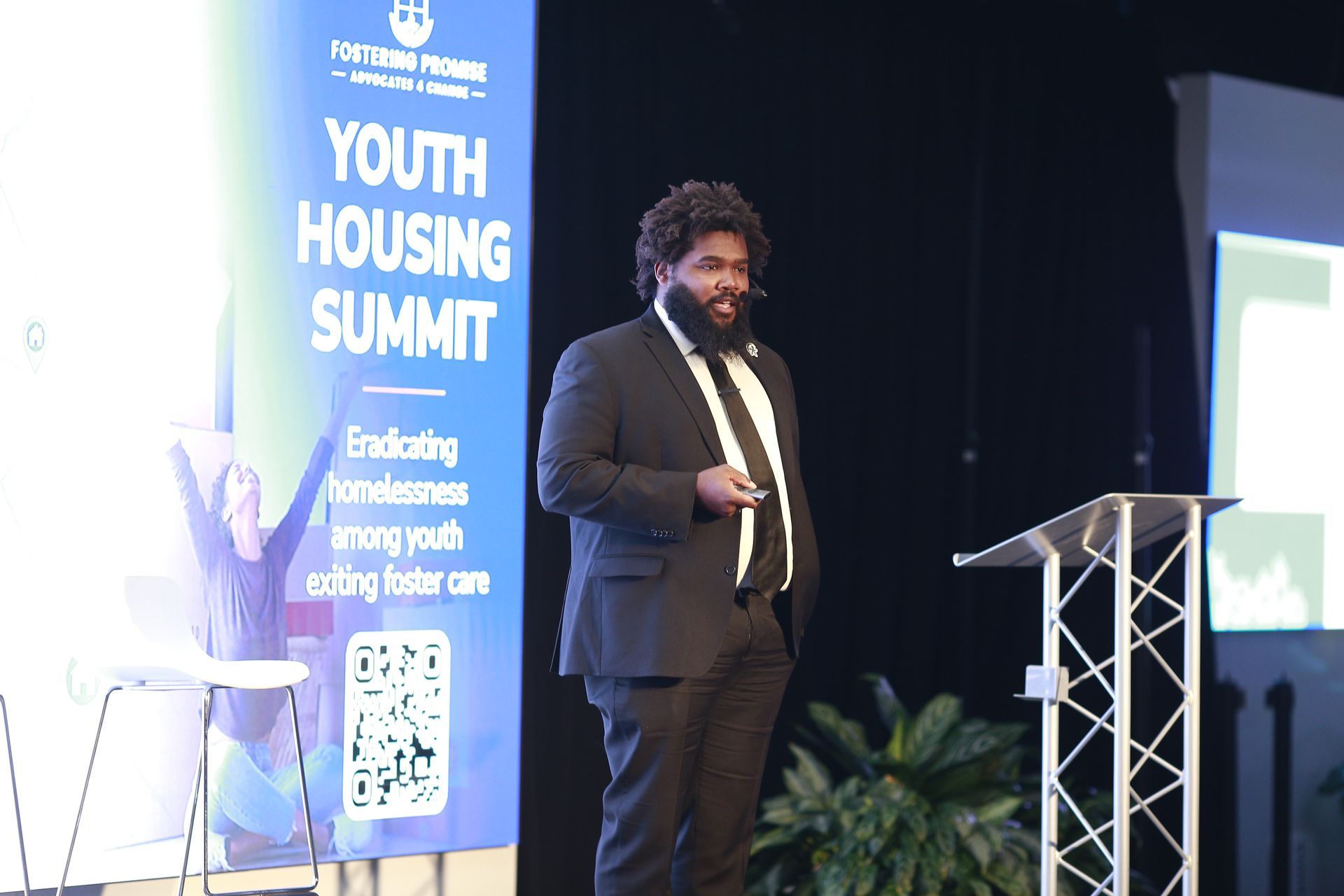Speaker at a podium during a Youth Housing Summit, with a blue event banner and stage backdrop.