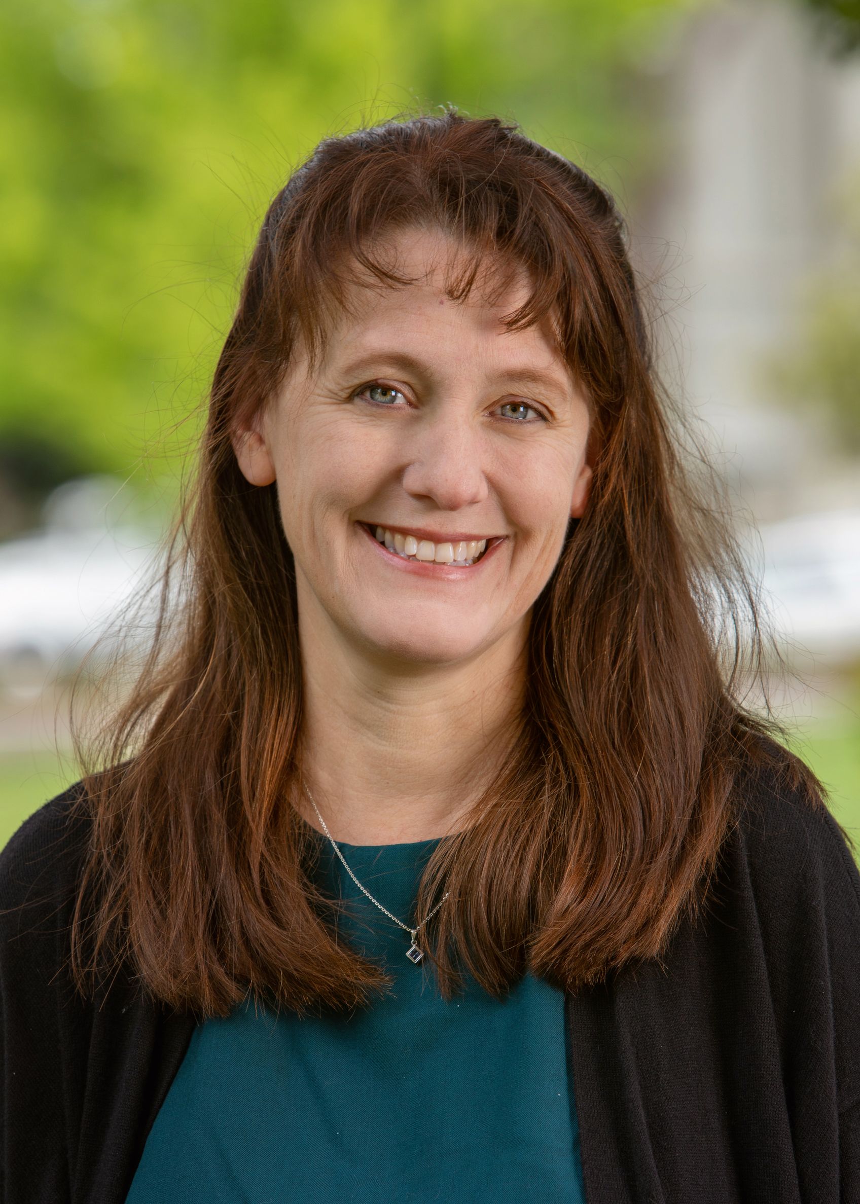 Smiling woman with long brown hair, wearing a teal top and black cardigan outdoors with a blurred green background