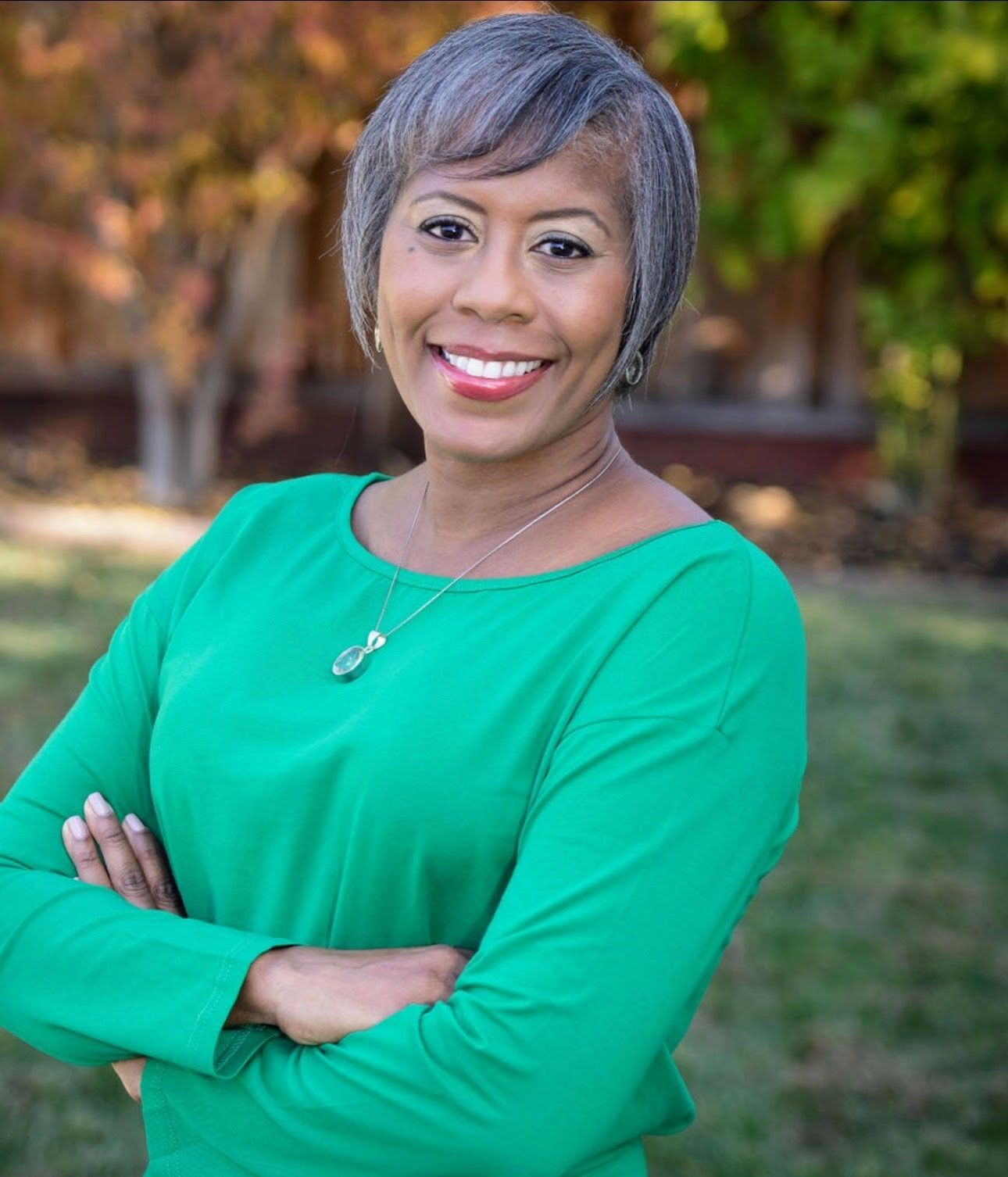 Smiling person in a bright green top with arms crossed, standing outdoors with blurred trees behind.