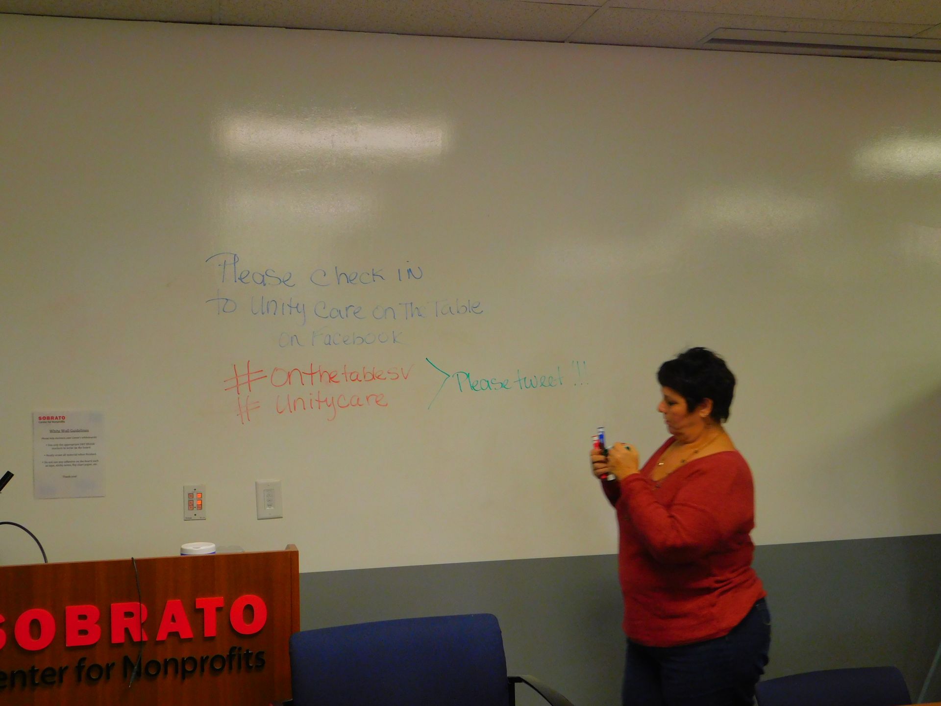 Person in red shirt speaking beside a whiteboard in a classroom or lecture room