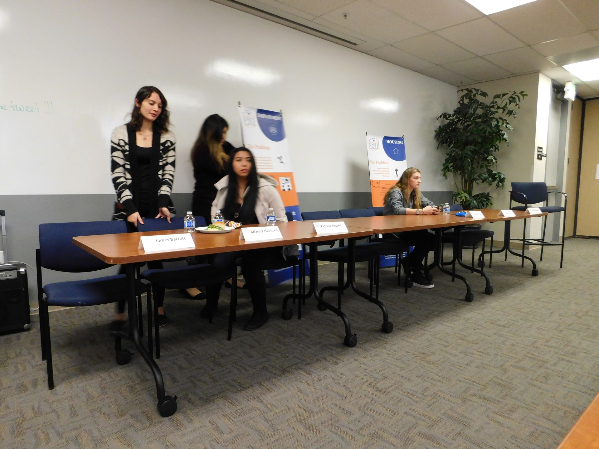 Three people sit and stand behind a long table in a conference room with name placards and posters.