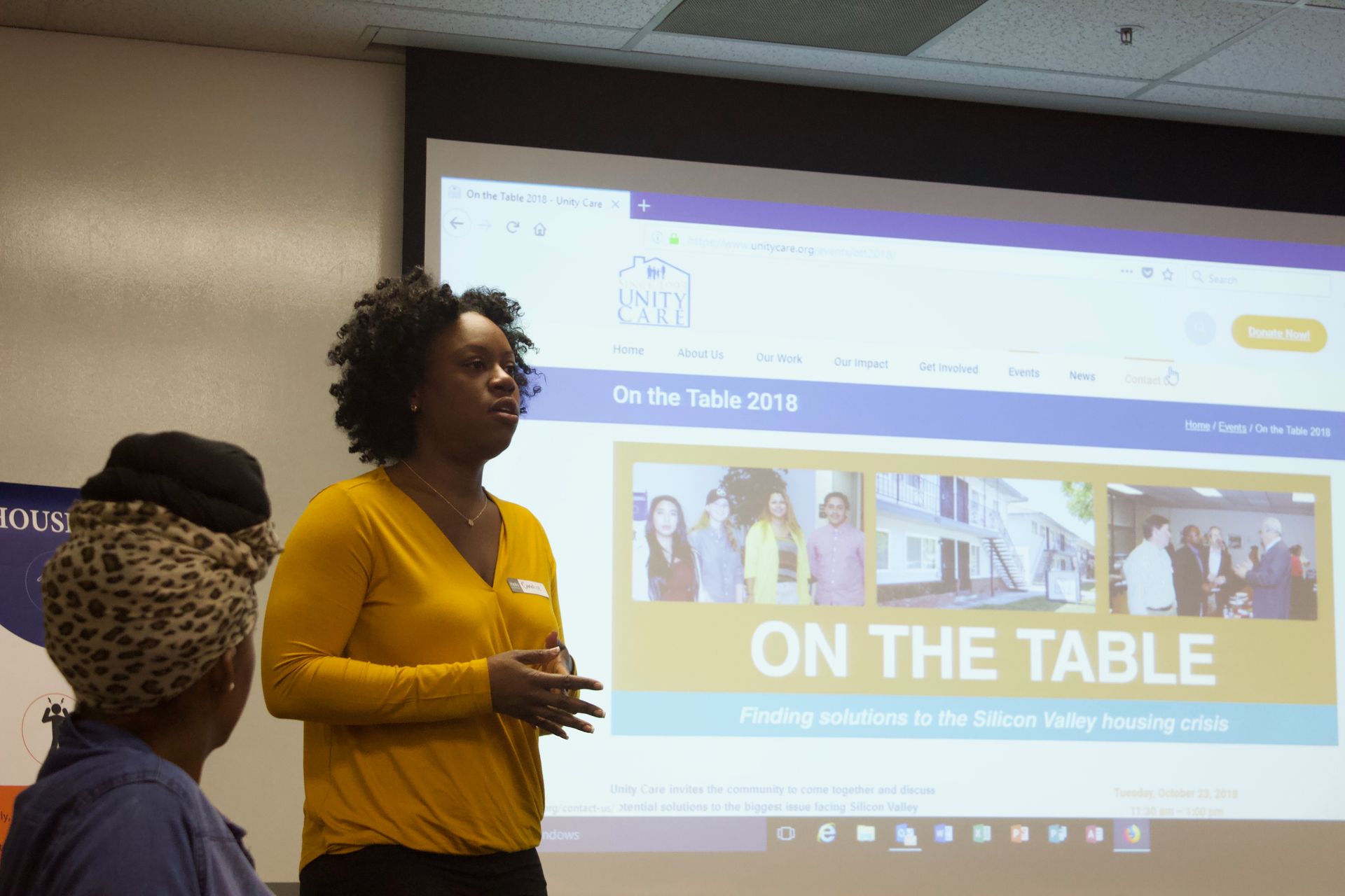 Woman in a yellow top presenting before a projected slide reading “ON THE TABLE”
