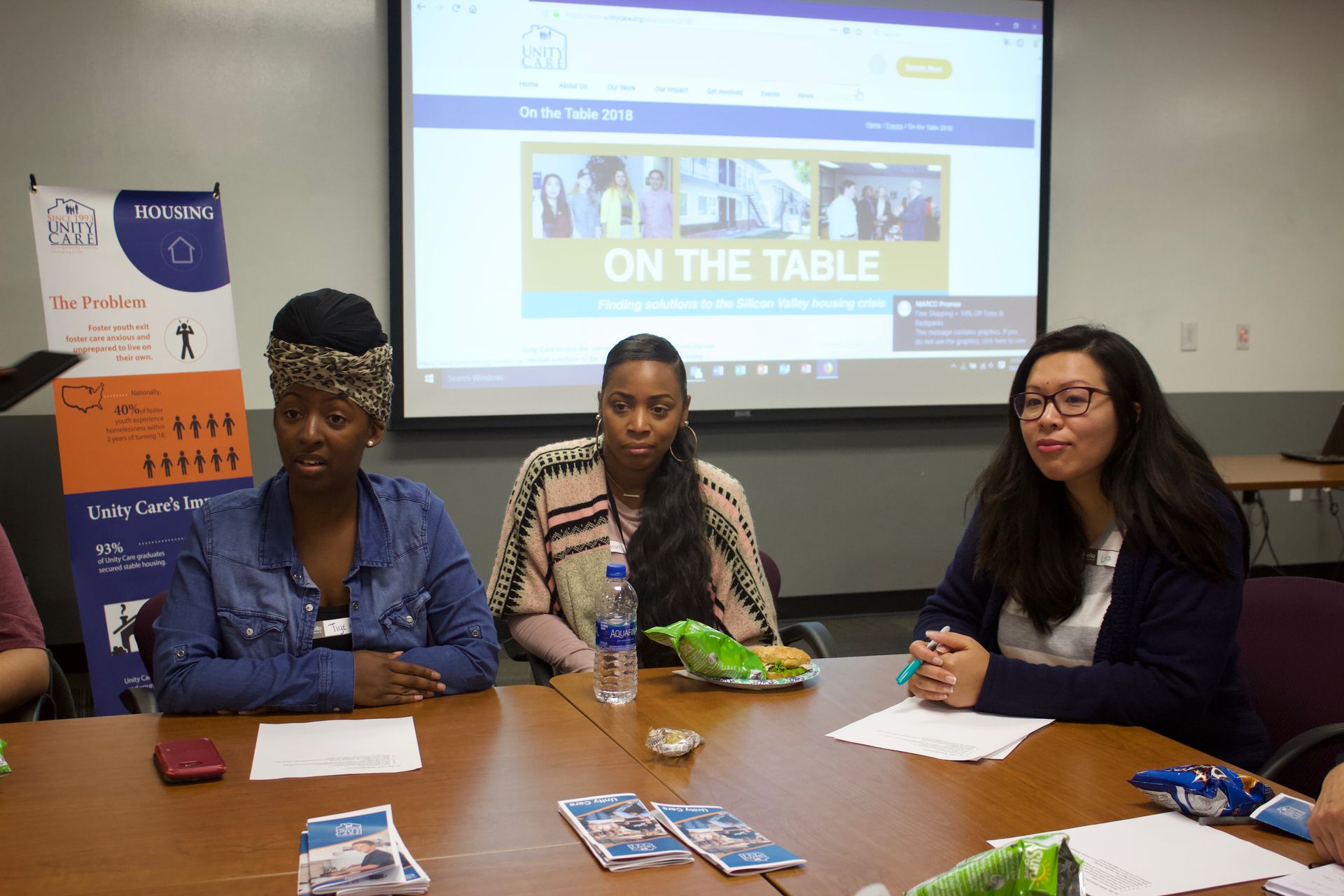 Three people seated at a table in a meeting room, with pamphlets and a projected presentation behind them.