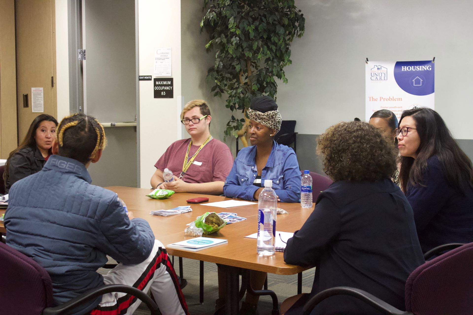 People seated around a table in a meeting room, discussing with papers, snacks, and water bottles.