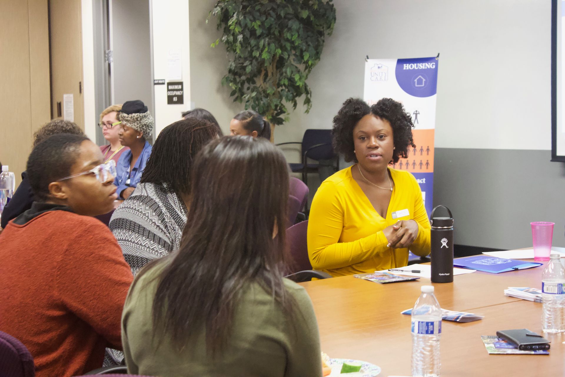 People seated around a conference table in a meeting room, with one woman in a yellow sweater speaking.