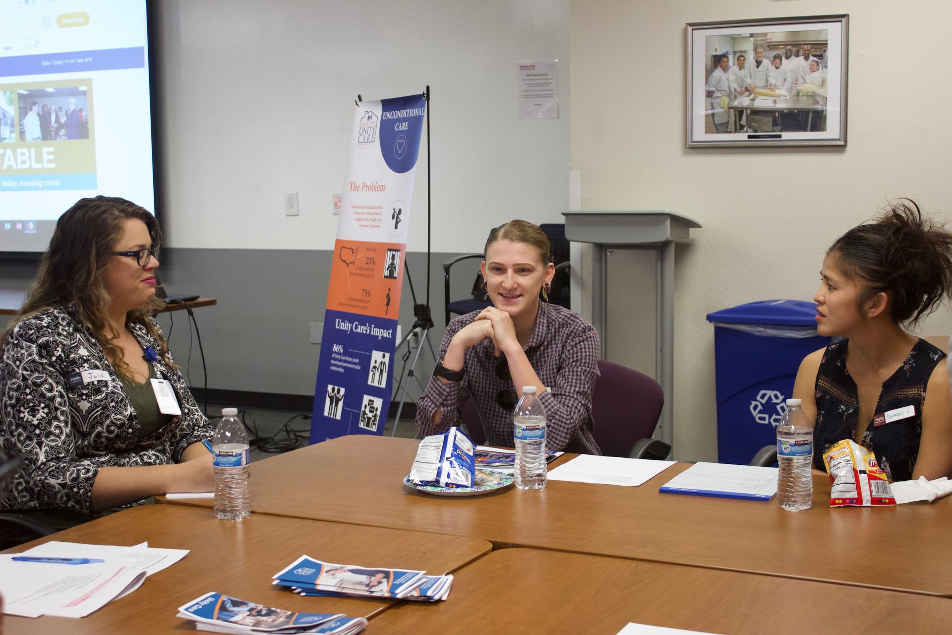 Three women seated at a conference table in a meeting room, talking with papers and water bottles.