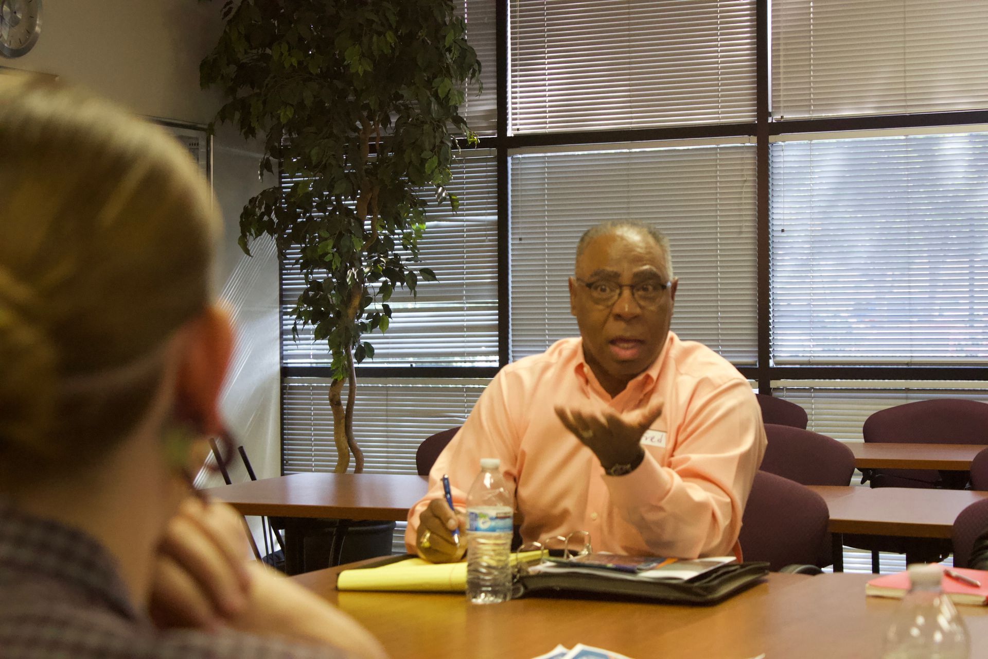 Man speaking at a table in a bright room, gesturing toward a listener in the foreground.