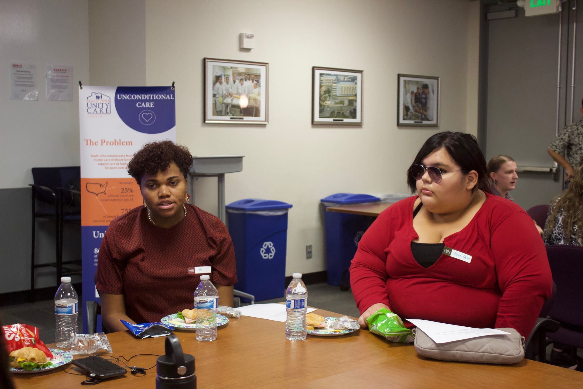 Two people seated at a table in a meeting room, with water bottles and snacks in front of them.