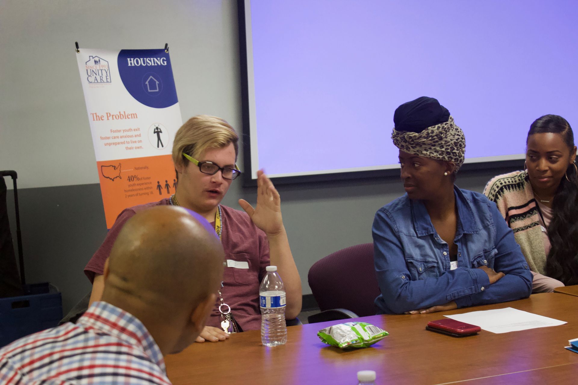Small group discussing at a conference table, with one person speaking beside a water bottle and snacks.