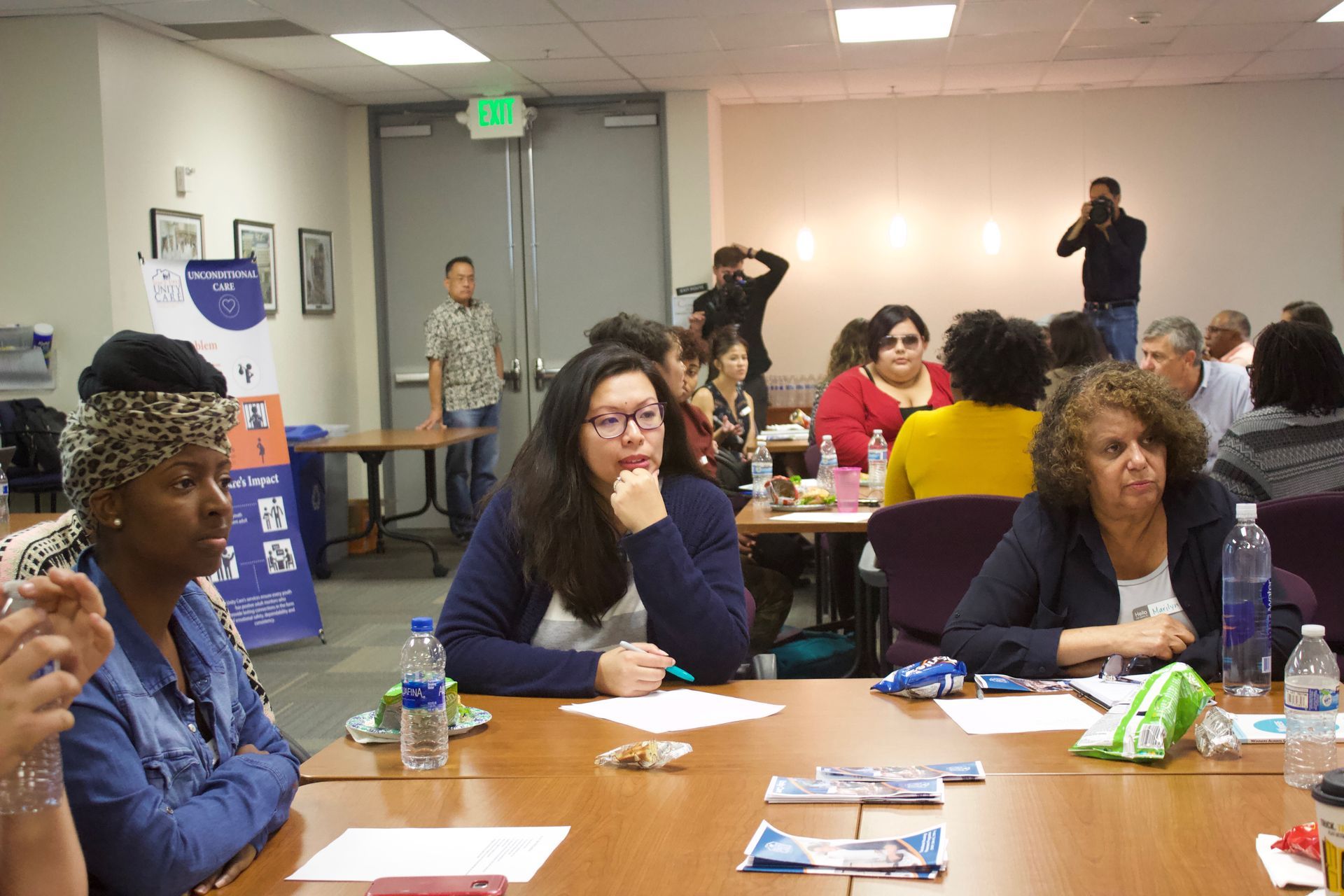 People seated at tables in a meeting room, listening and taking notes.